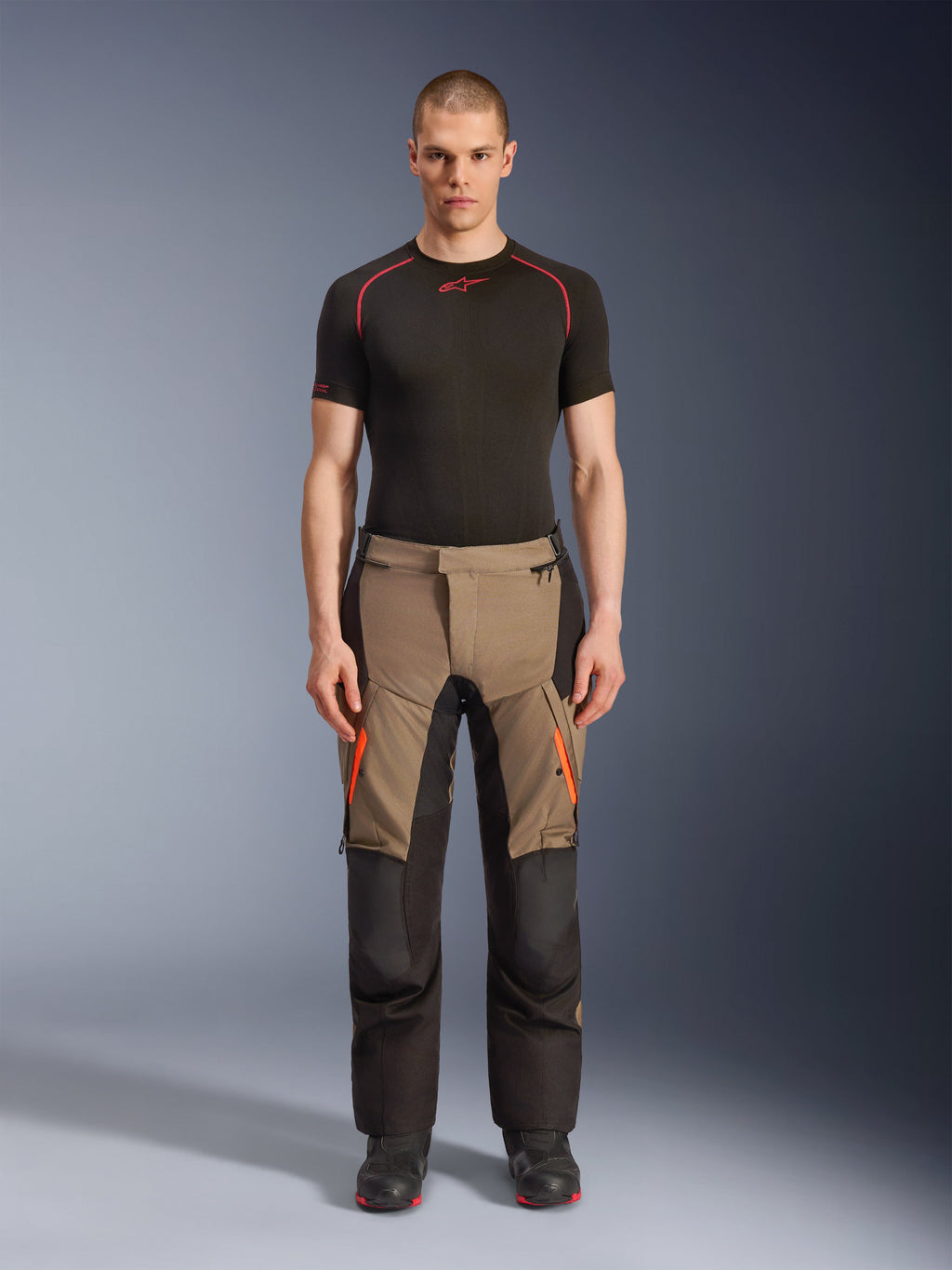 Mannequin homme portant le pantalon Alpinestars Andes V4 Drystar® en Walnut Black, pantalon de moto d'aventure associé à une couche de base noire et des bottes, avec des accents de fermeture éclair orange sur les cuisses et des panneaux noirs renforcés pour une durabilité tout-terrain.