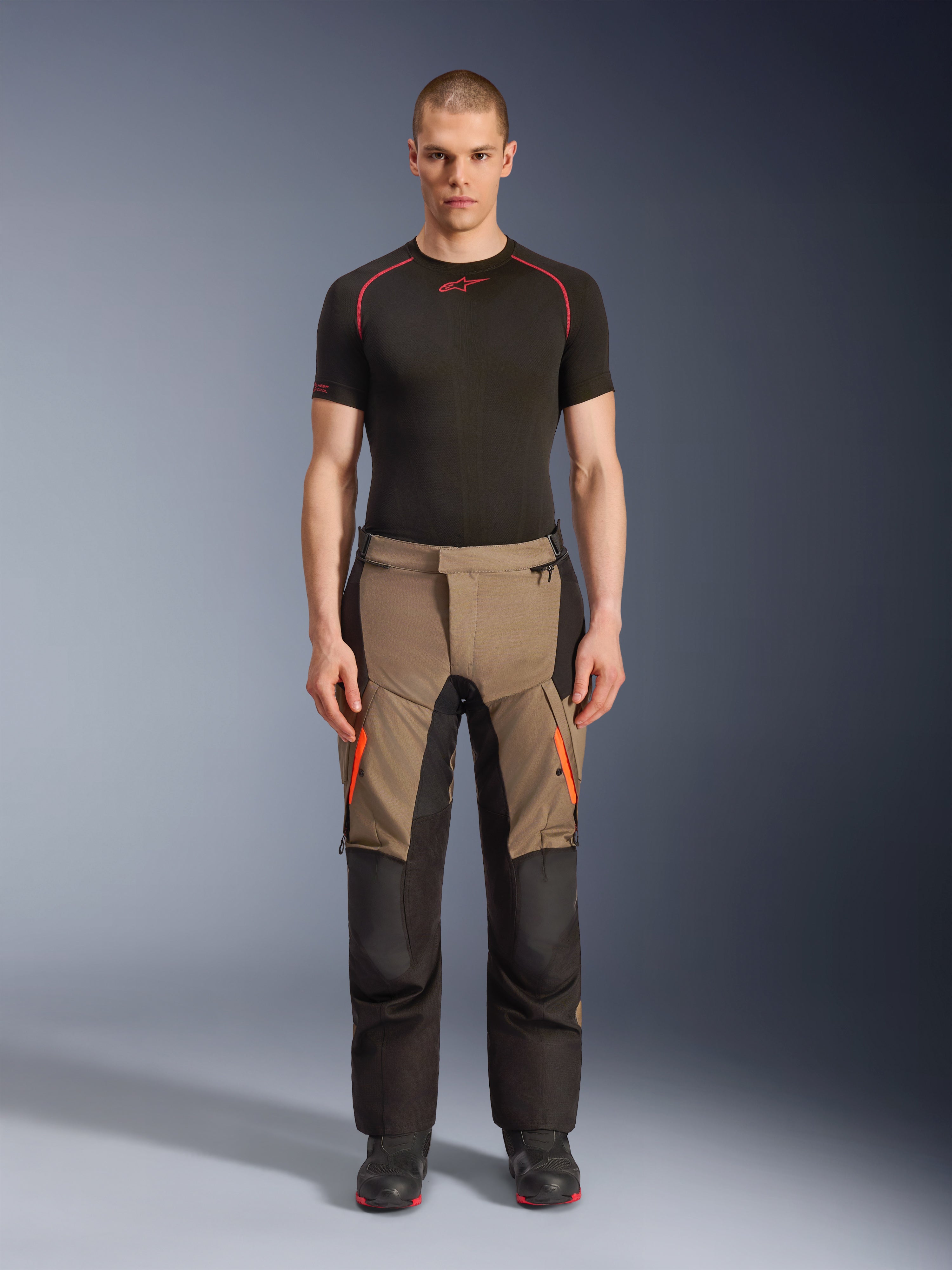Mannequin homme portant le pantalon Alpinestars Andes V4 Drystar® en Walnut Black, pantalon de moto d'aventure associé à une couche de base noire et des bottes, avec des accents de fermeture éclair orange sur les cuisses et des panneaux noirs renforcés pour une durabilité tout-terrain.