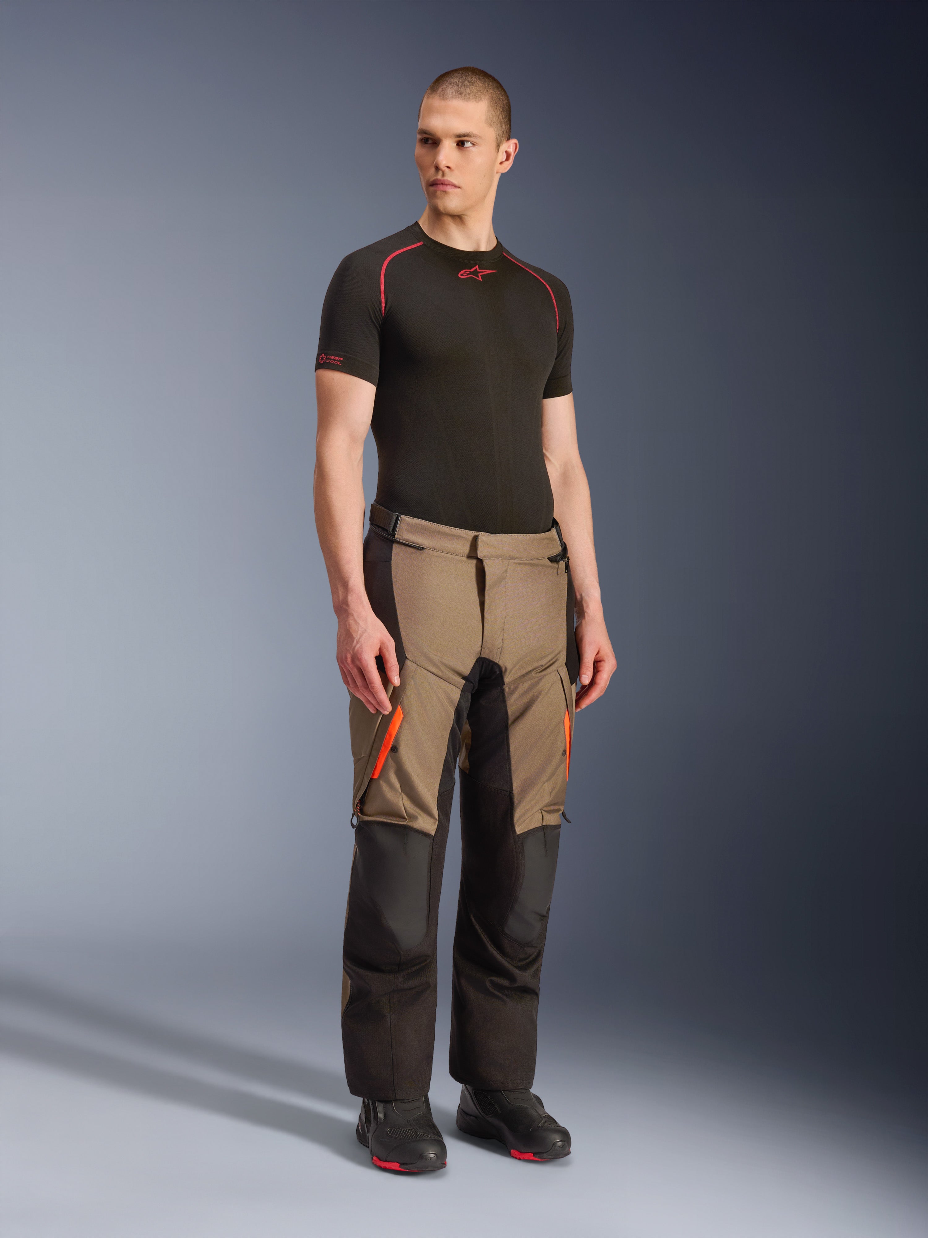 Pantalon Alpinestars Andes V4 Drystar®, pantalon de moto aventure touring, Walnut Black avec accents orange, porté par un mannequin avec un haut technique noir à manches courtes et des bottes noires, doté de panneaux textiles renforcés et de poches cargo, conception robuste pour rouler par tous les temps
