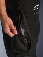 Pantalon Alpinestars Andes V4 Drystar, noir, gros plan sur l'ouverture d'une aération de cuisse par une main, doublure en mesh pour la circulation de l'air, détail de fermeture éclair étanche, logo Alpinestars blanc