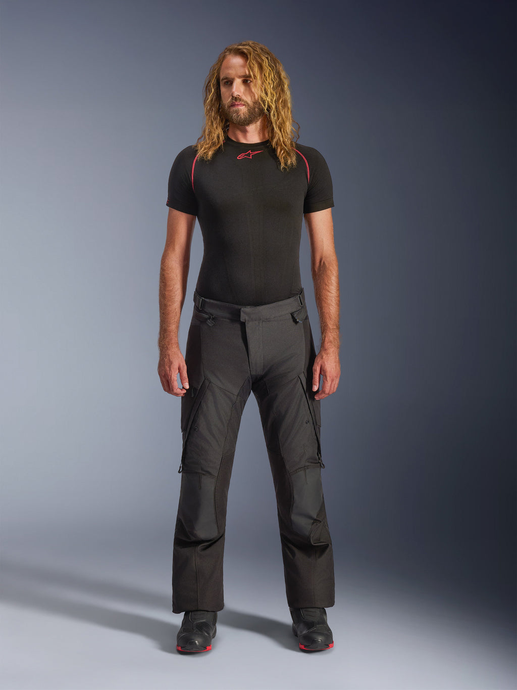Mannequin homme aux cheveux longs portant le pantalon Alpinestars Andes V4 Drystar® noir, pantalon de moto adventure touring avec panneaux de genoux renforcés et poches cargo, associé à une couche de base noire à manches courtes et des bottes de moto.