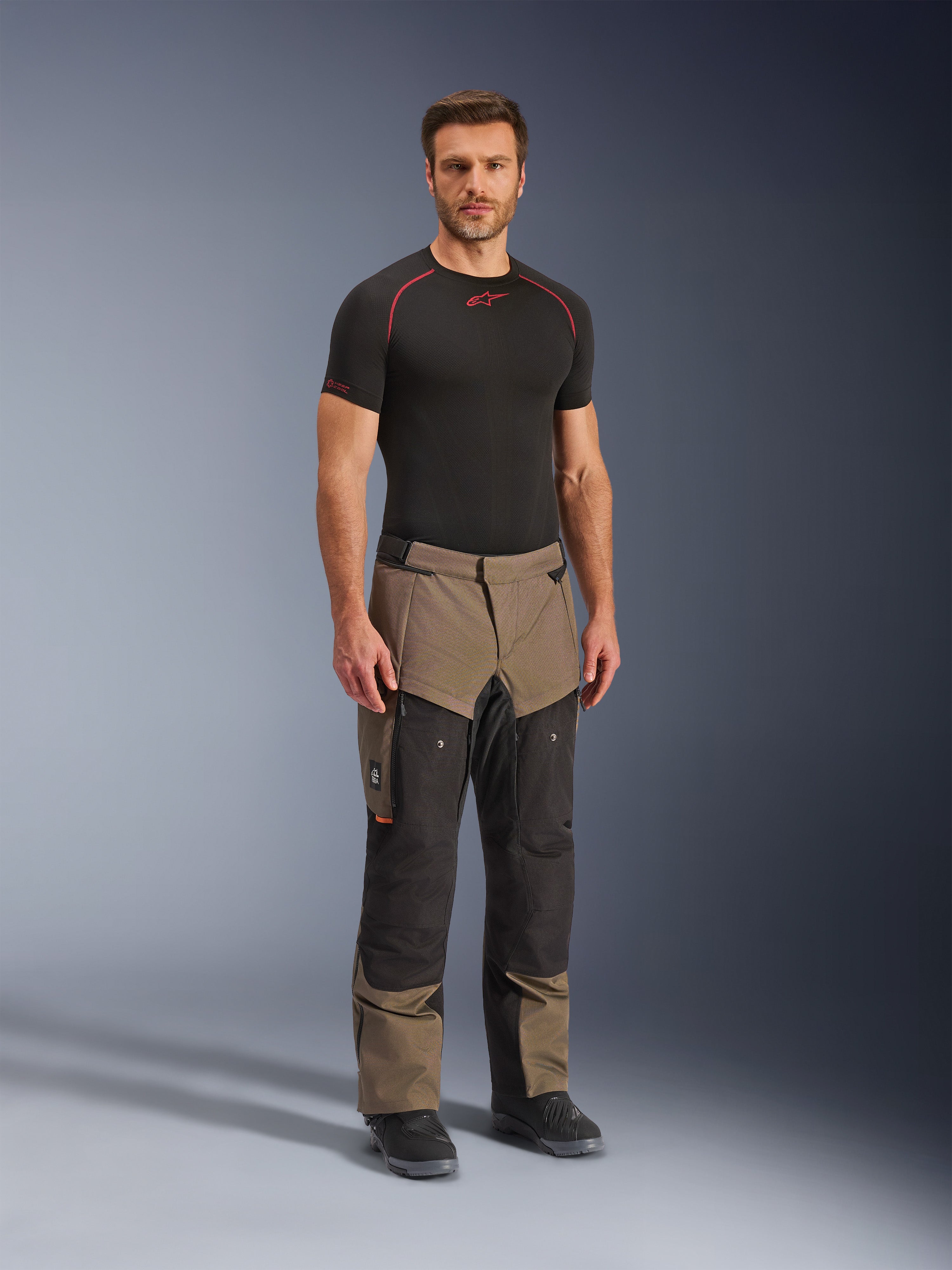 Pantalon Alpinestars Cusco Drystar®, pantalon de moto aventure, noyer et noir, porté par un mannequin avec une couche de base noire et des bottes, doté de grandes poches cargo sur les cuisses, de fermetures éclair de ventilation et d'un design bicolore robuste.