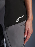 Gros plan sur le pantalon Alpinestars Stella Andes V4 Drystar® pour femme en noir et gris foncé, présentant un logo étoile blanc sur un empiècement noir et le marquage Alpinestars noir sur un empiècement gris, montrant le textile technique et le détail d'une poche zippée.