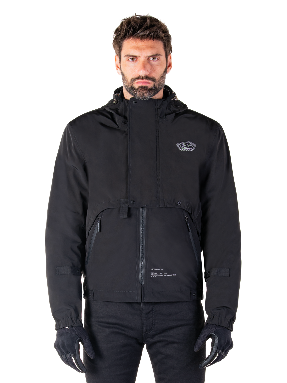 Alpinestars Mootant Waterproof Parka, schwarze Motorradjacke mit Kapuze und modularem technischem Design, getragen von einem männlichen Model mit schwarzen Handschuhen, mit einer einzigartigen Brustklappe und Seitentaschen mit Reißverschluss, urbaner und Touring-Fahrstil.