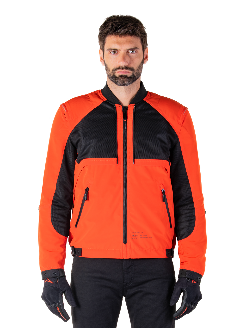 Parka imperméable Mootant Alpinestars en Spicy Orange et noir, veste de moto modulaire présentée dans une configuration de style bomber sur un mannequin homme, avec des empiècements en maille noire sur le haut de la poitrine, le buste et les manches orange vif, une fermeture éclair frontale et un col côtelé, portée avec des gants et un pantalon noirs.