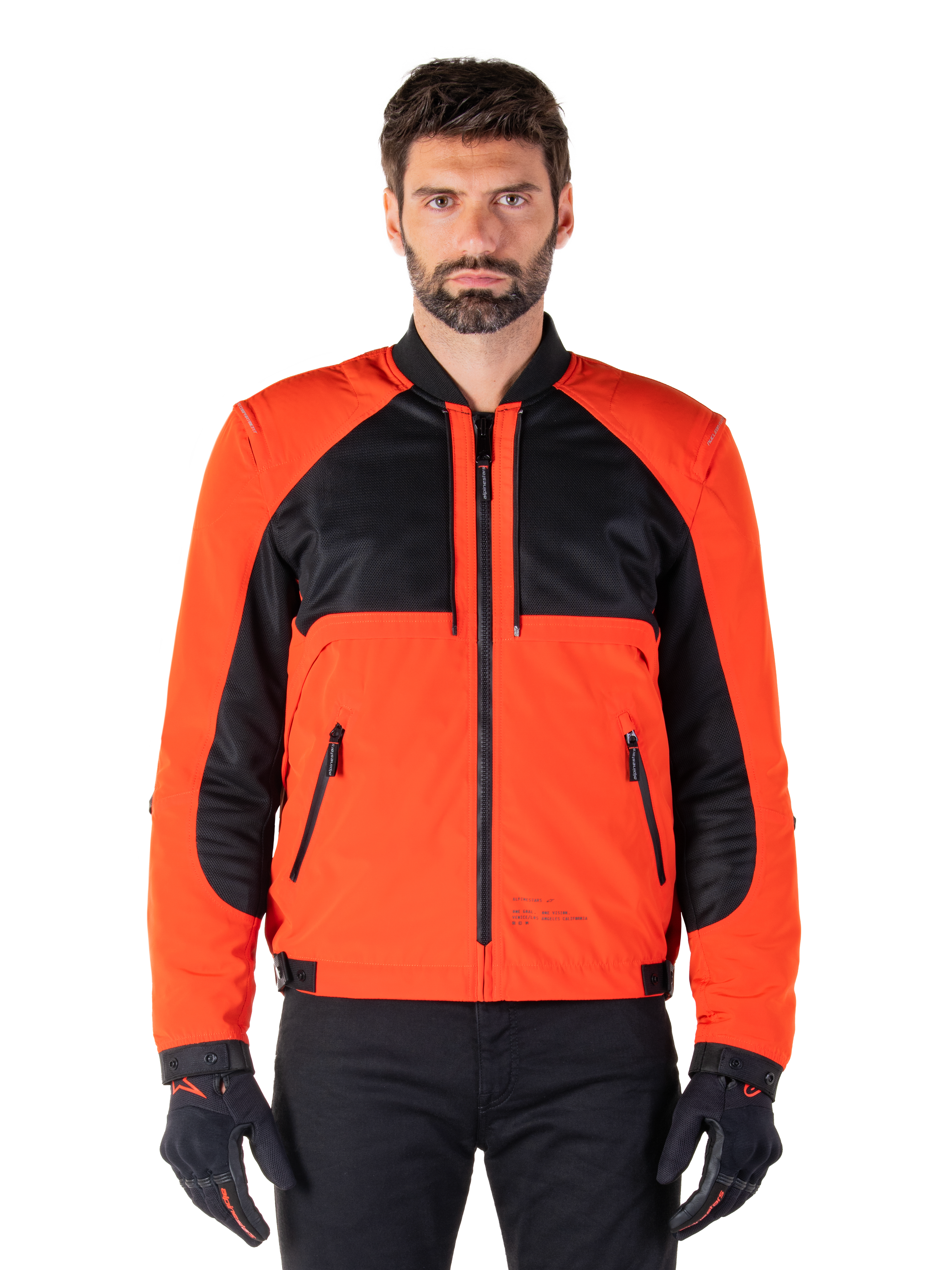 Parka imperméable Mootant Alpinestars en Spicy Orange et noir, veste de moto modulaire présentée dans une configuration de style bomber sur un mannequin homme, avec des empiècements en maille noire sur le haut de la poitrine, le buste et les manches orange vif, une fermeture éclair frontale et un col côtelé, portée avec des gants et un pantalon noirs.