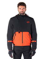 Alpinestars Mootant Waterproof Parka, veste de moto modulaire en Spicy Orange et noir, portée par un mannequin avec des gants assortis, présente un design à capuche avec des empiècements bicolores, une fermeture éclair frontale et des poignets réglables, vêtement de moto urbain contemporain