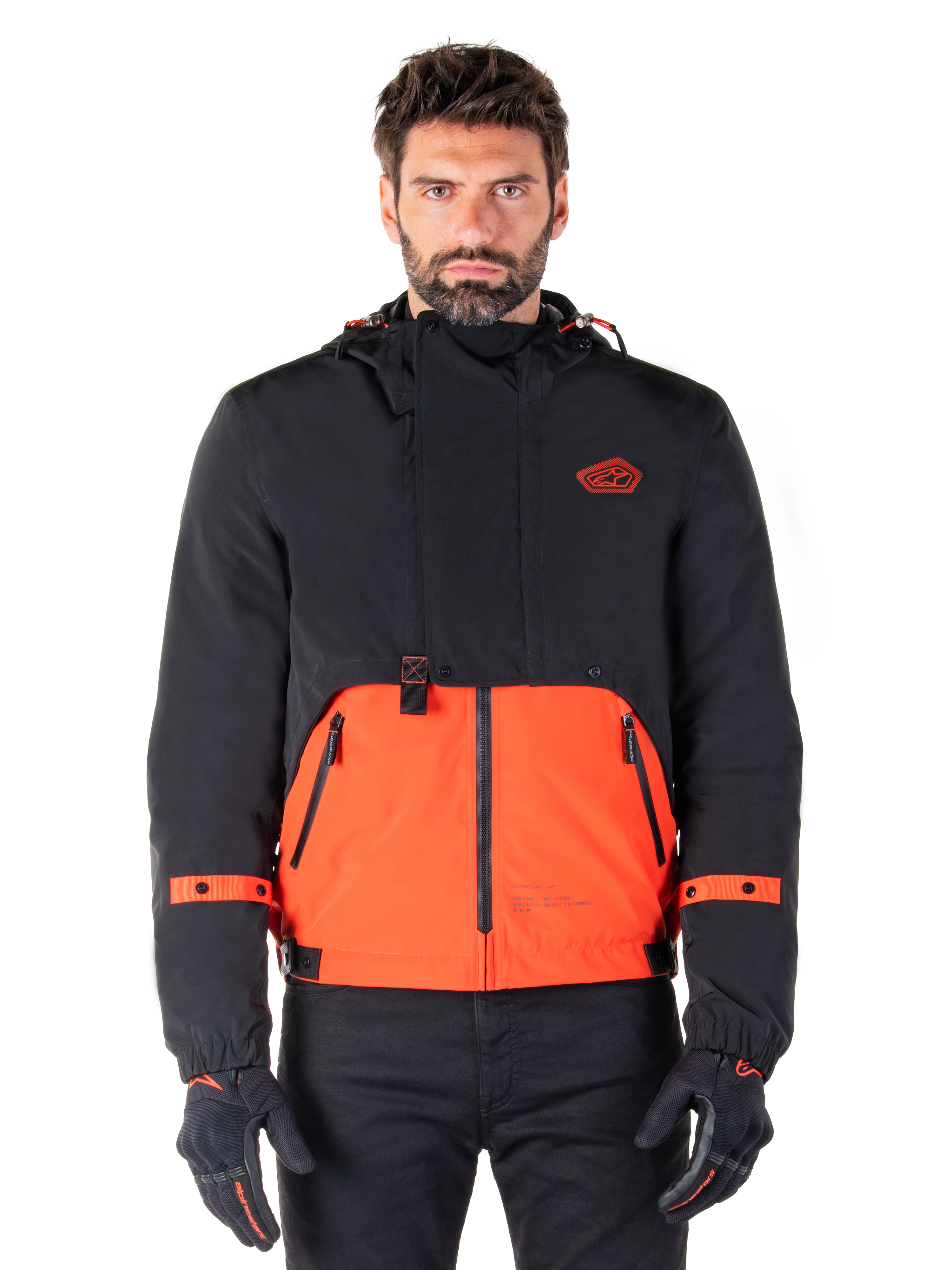 Alpinestars Mootant Waterproof Parka, modulaire motorjas in Spicy Orange en zwart, gedragen door een model met bijpassende handschoenen, voorzien van een ontwerp met capuchon en tweekleurige panelen, ritssluiting aan de voorzijde en verstelbare manchetten, urban contemporary motorkleding