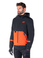 Homme portant la Parka Alpinestars Mootant Waterproof en Spicy Orange et noir, une veste de moto modulaire avec une couche supérieure noire à capuche et une section inférieure orange vif, accompagnée de gants et d'un pantalon de moto noirs.