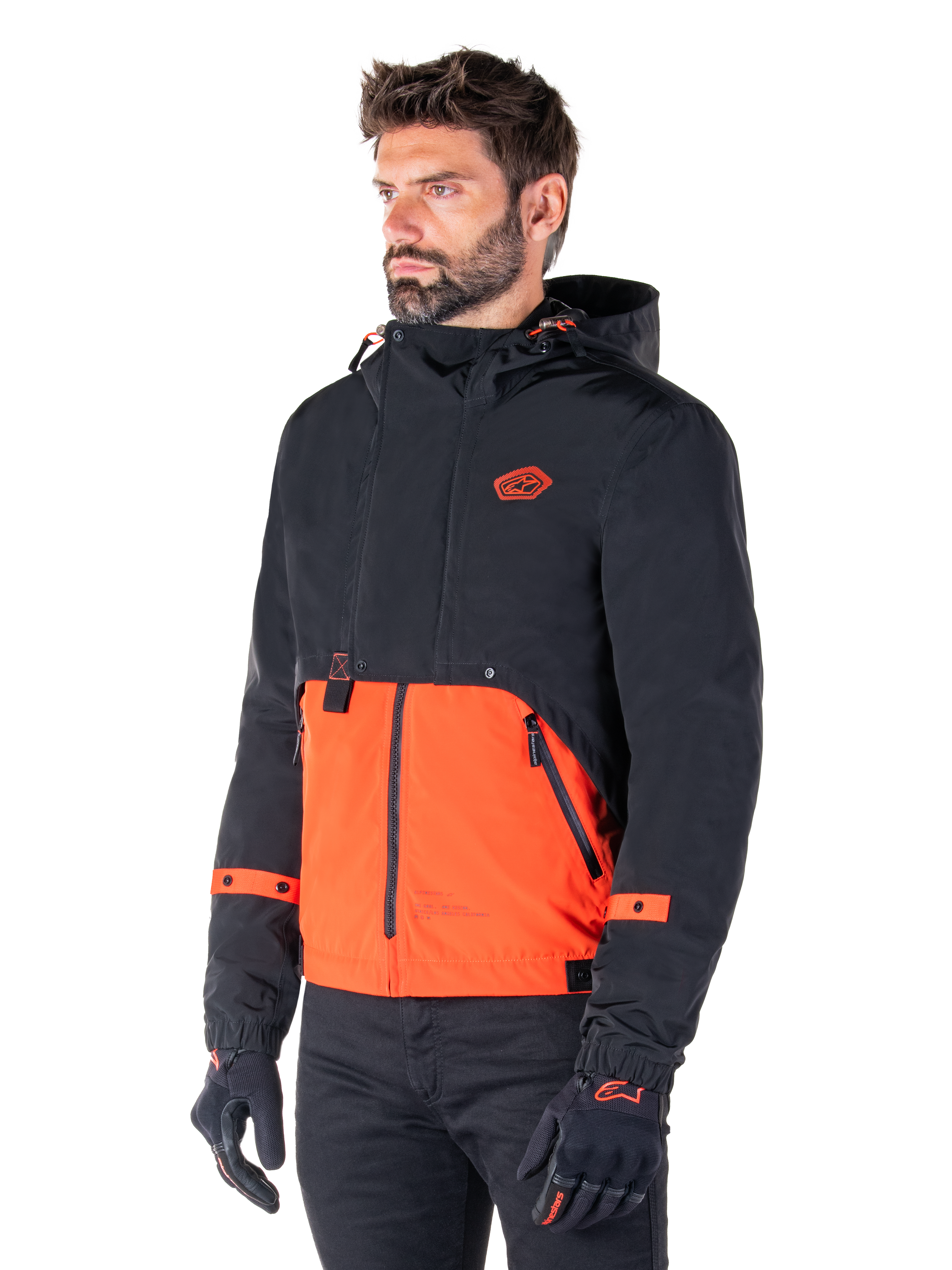 Homme portant la Parka Alpinestars Mootant Waterproof en Spicy Orange et noir, une veste de moto modulaire avec une couche supérieure noire à capuche et une section inférieure orange vif, accompagnée de gants et d'un pantalon de moto noirs.