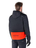 Parka imperméable Alpinestars Mootant, orange épicé et noir, vue arrière sur un mannequin homme, dotée d'une partie supérieure noire à capuche et d'une partie inférieure orange vif avec le logo MO.ST.EQ., veste de moto urbaine contemporaine portée avec un pantalon et des gants noirs