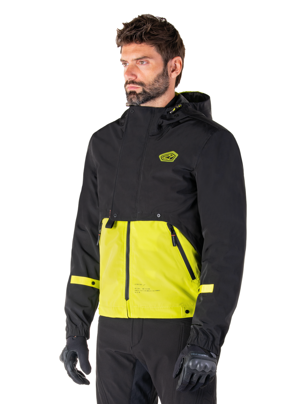 Alpinestars Mootant Waterproof Parka in Lime Punch Black, modulaire motorjas gedragen door een man met zwarte motorhandschoenen en broek, met een zwart bovenstuk en mouwen en een opvallend limoengroen paneel aan de onderkant, ontwerp met capuchon en een utilitaire, militair geïnspireerde esthetiek