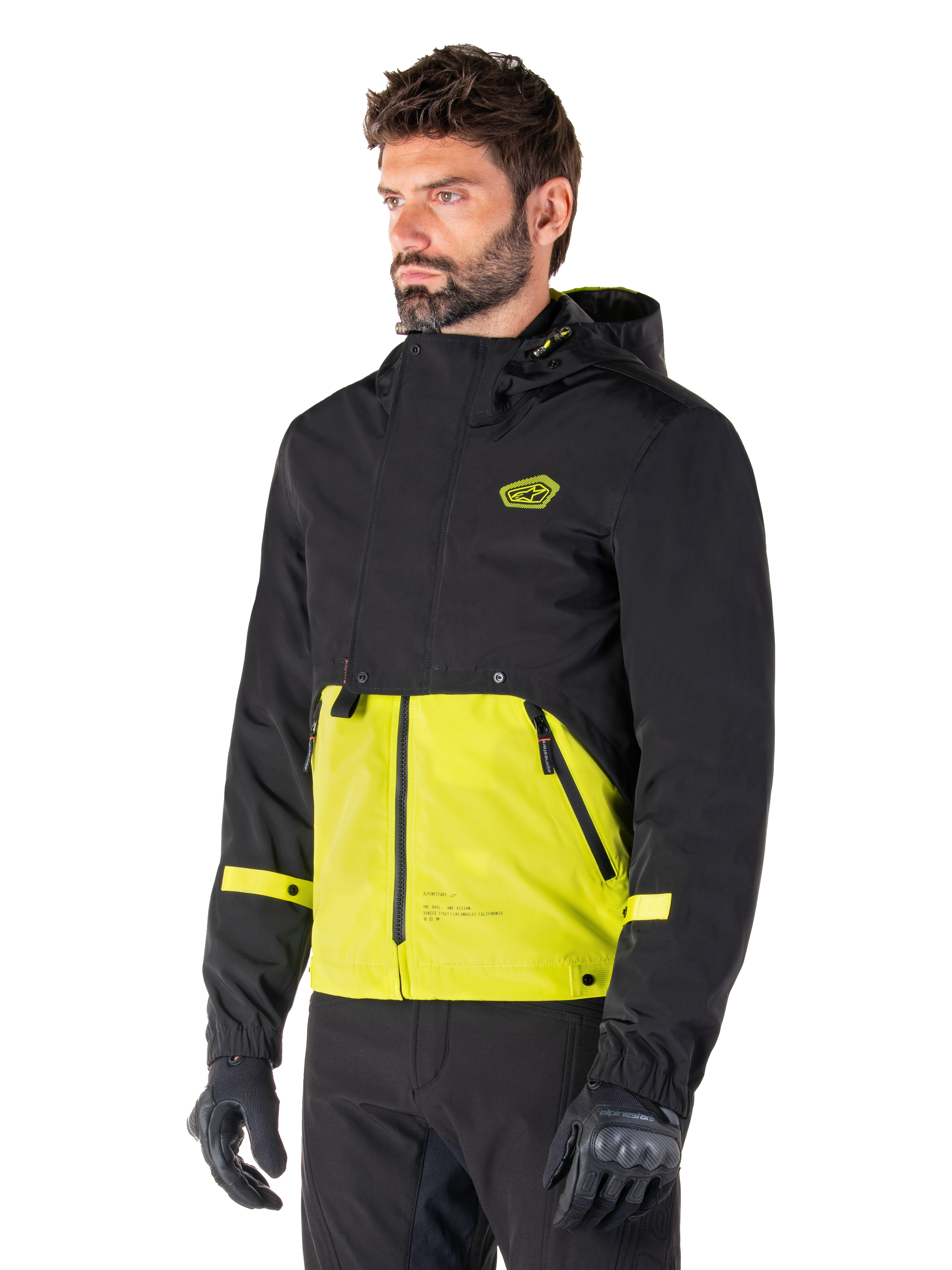 Alpinestars Mootant Waterproof Parka in Lime Punch Black, modulaire motorjas gedragen door een man met zwarte motorhandschoenen en broek, met een zwart bovenstuk en mouwen en een opvallend limoengroen paneel aan de onderkant, ontwerp met capuchon en een utilitaire, militair geïnspireerde esthetiek