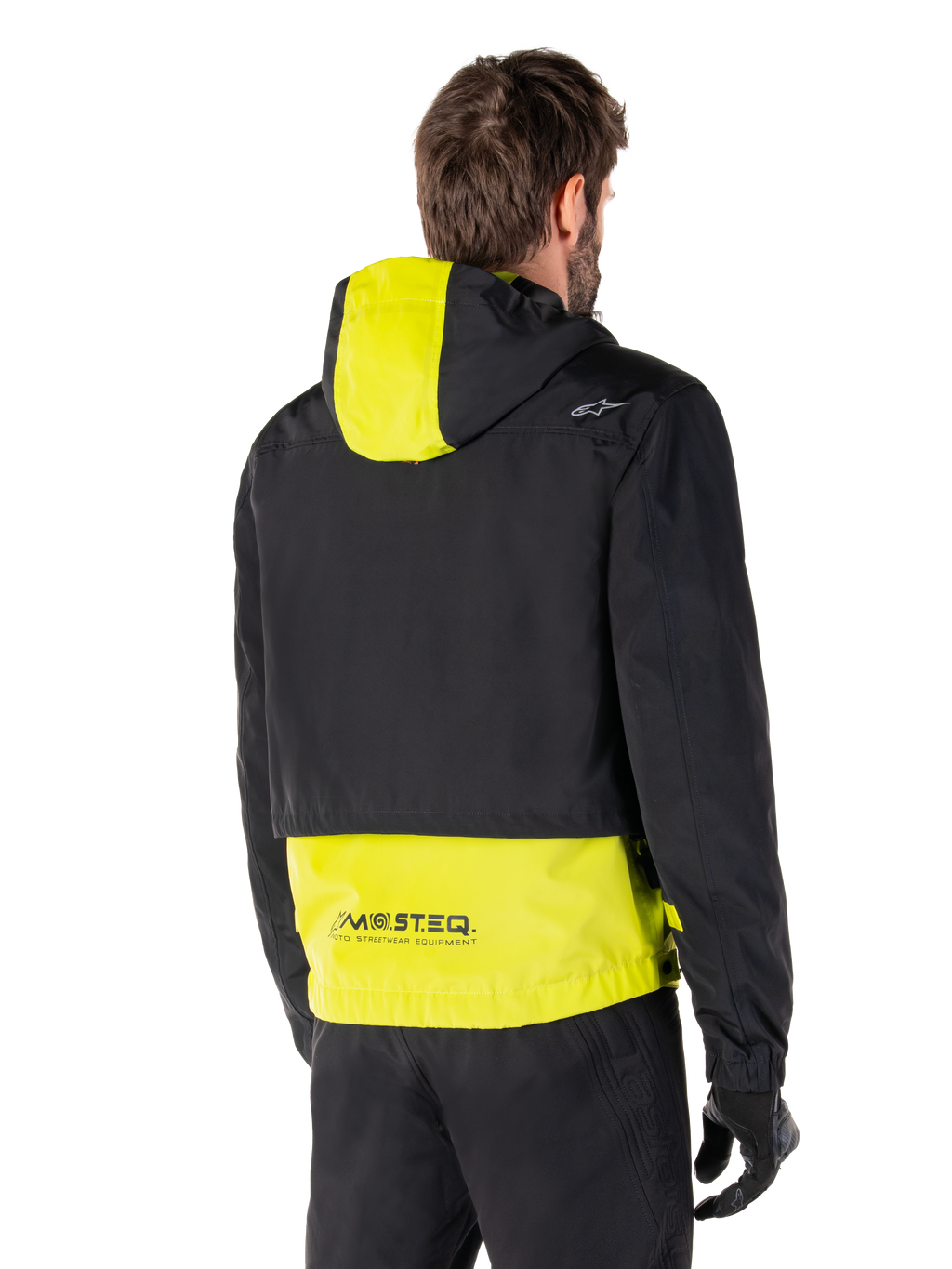 Alpinestars Mootant Waterproof Parka, achteraanzicht, Lime Punch en zwart, zwart bovendeel met een limoengeel paneel op de capuchon en de onderrug, MO.ST.EQ. branding op de zoom, gedragen met een zwarte motorbroek en handschoenen, urban contemporary motorjas