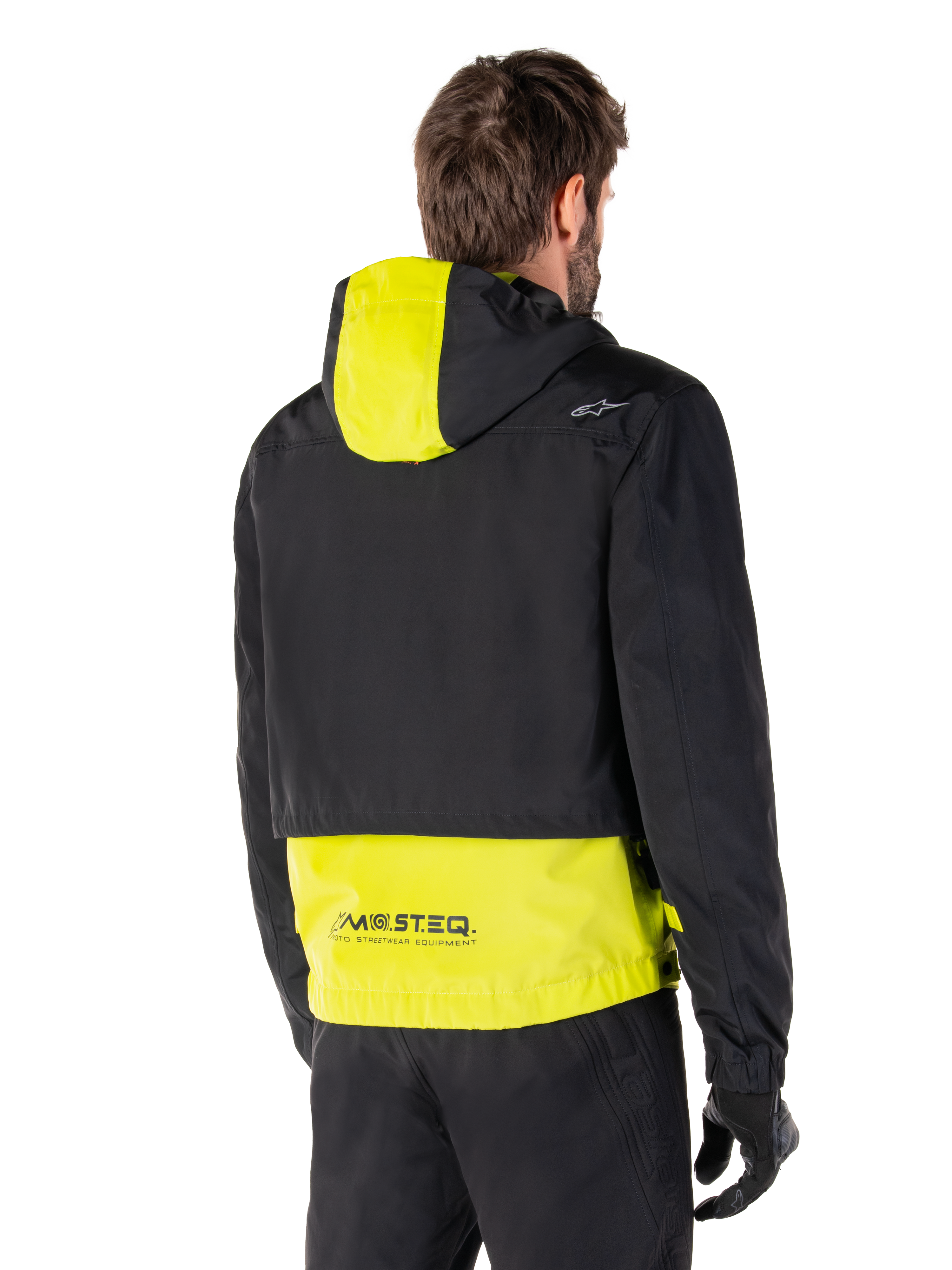 Alpinestars Mootant Waterproof Parka, achteraanzicht, Lime Punch en zwart, zwart bovendeel met een limoengeel paneel op de capuchon en de onderrug, MO.ST.EQ. branding op de zoom, gedragen met een zwarte motorbroek en handschoenen, urban contemporary motorjas
