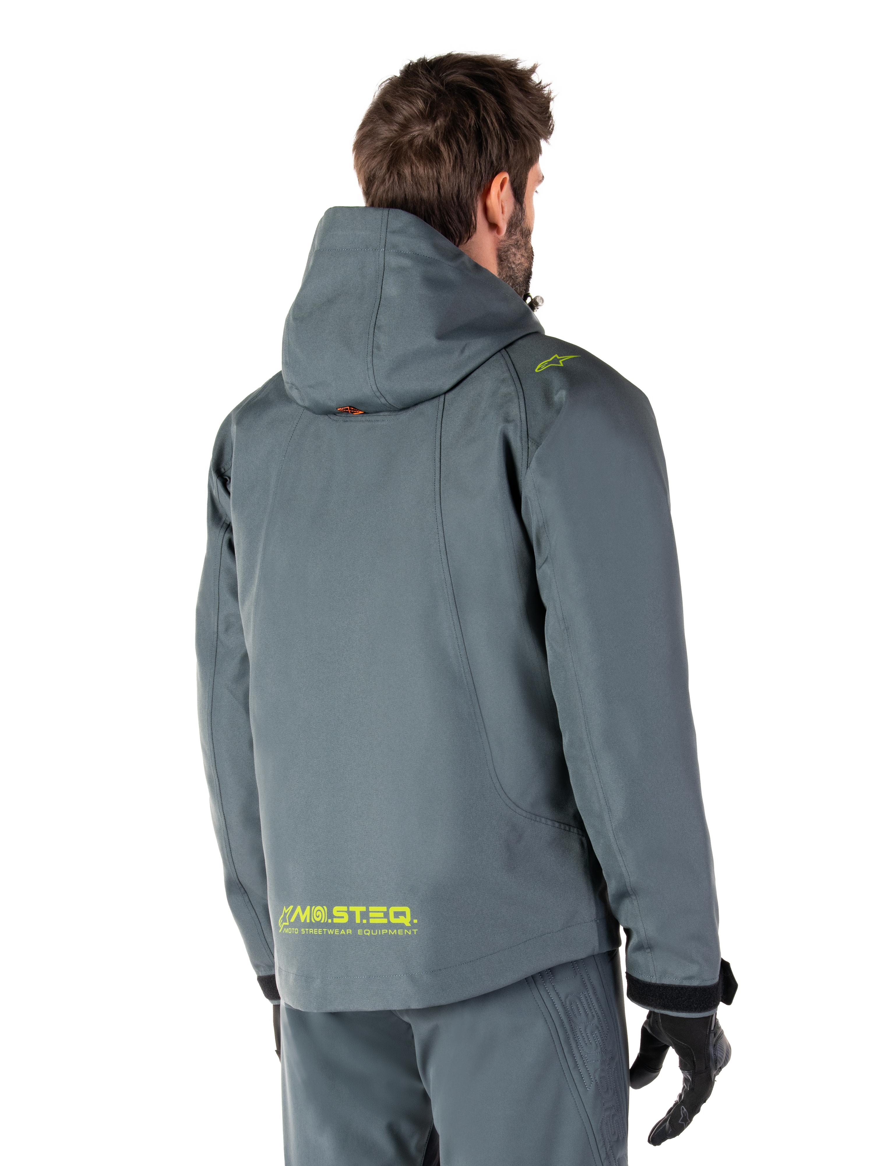 Parka imperméable Alpinestars Morush en Urban Gray, vue de dos, avec marquage MO.ST.EQ. jaune sur le bas du dos et l'épaule, design minimaliste à capuche, portée avec un pantalon de moto gris assorti et des gants noirs, style streetwear moto urbain
