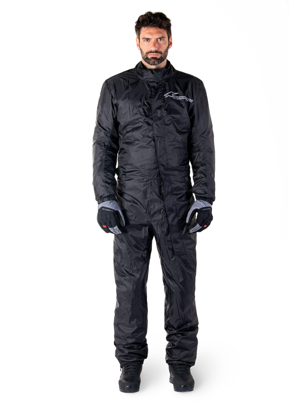 Alpinestars Hurricane Rain V2 Suit, volledig motorregenpak in het zwart, waterdichte beschermende kleding met hoge kraag en stormsluiting aan de voorzijde, voorzien van een wit logo op de borst, gedragen met zwart-grijze handschoenen en laarzen