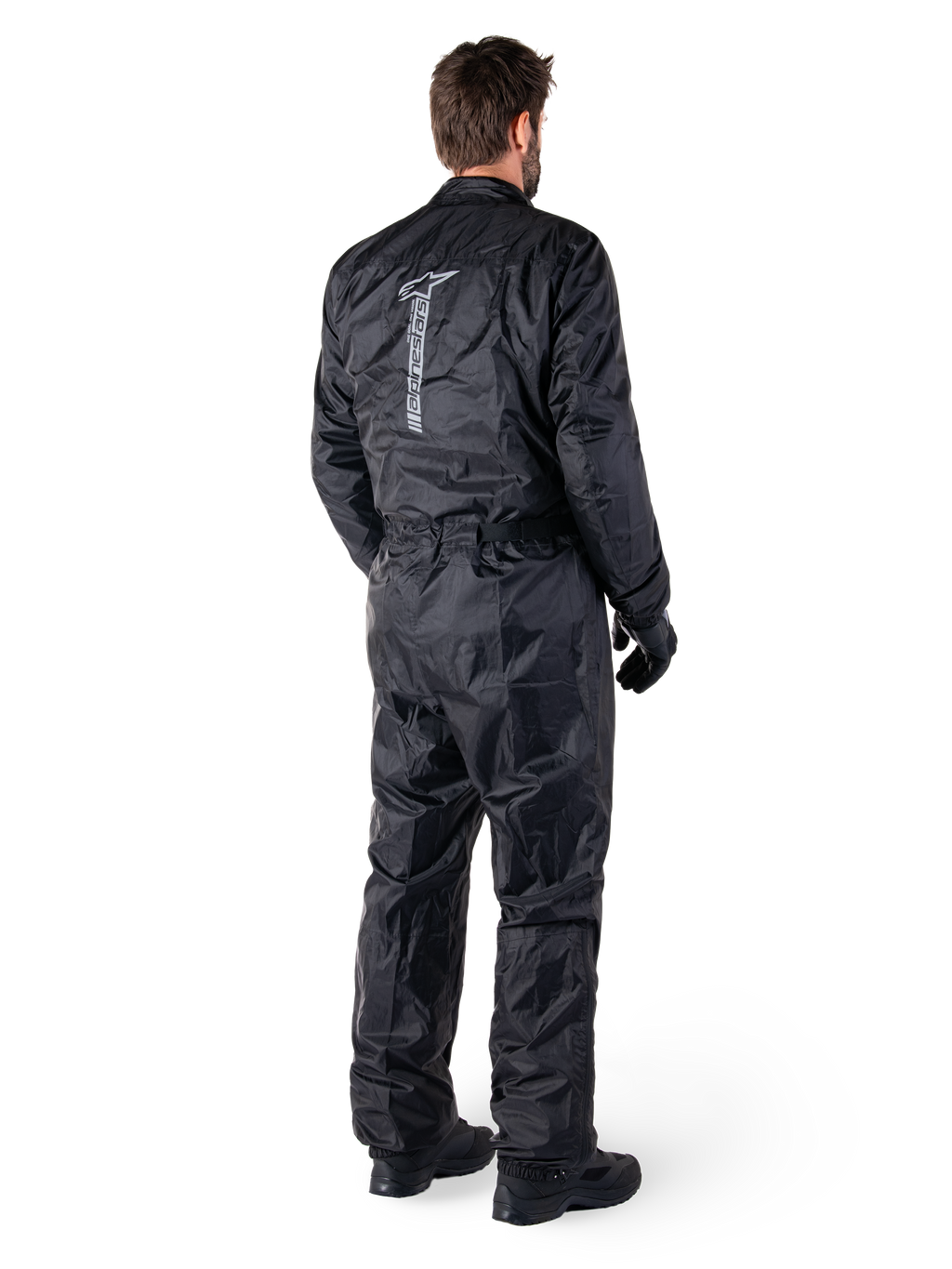 Alpinestars Hurricane Rain V2-pak, zwart eendelig waterdicht motorregenpak, achteraanzicht met groot verticaal reflecterend logo, lichtgewicht windbestendige stof, gedragen met zwarte handschoenen en laarzen