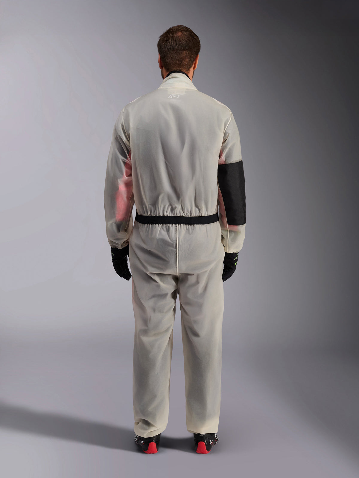 Alpinestars Kart Rain Suit, sur-combinaison de karting, transparente translucide, vue arrière montrant un panneau noir renforcé sur le coude droit pour la protection thermique, taille élastiquée avec bordure noire, matériau transparent laissant apparaître une combinaison de course en dessous, associée à des gants noirs et des bottines de course.