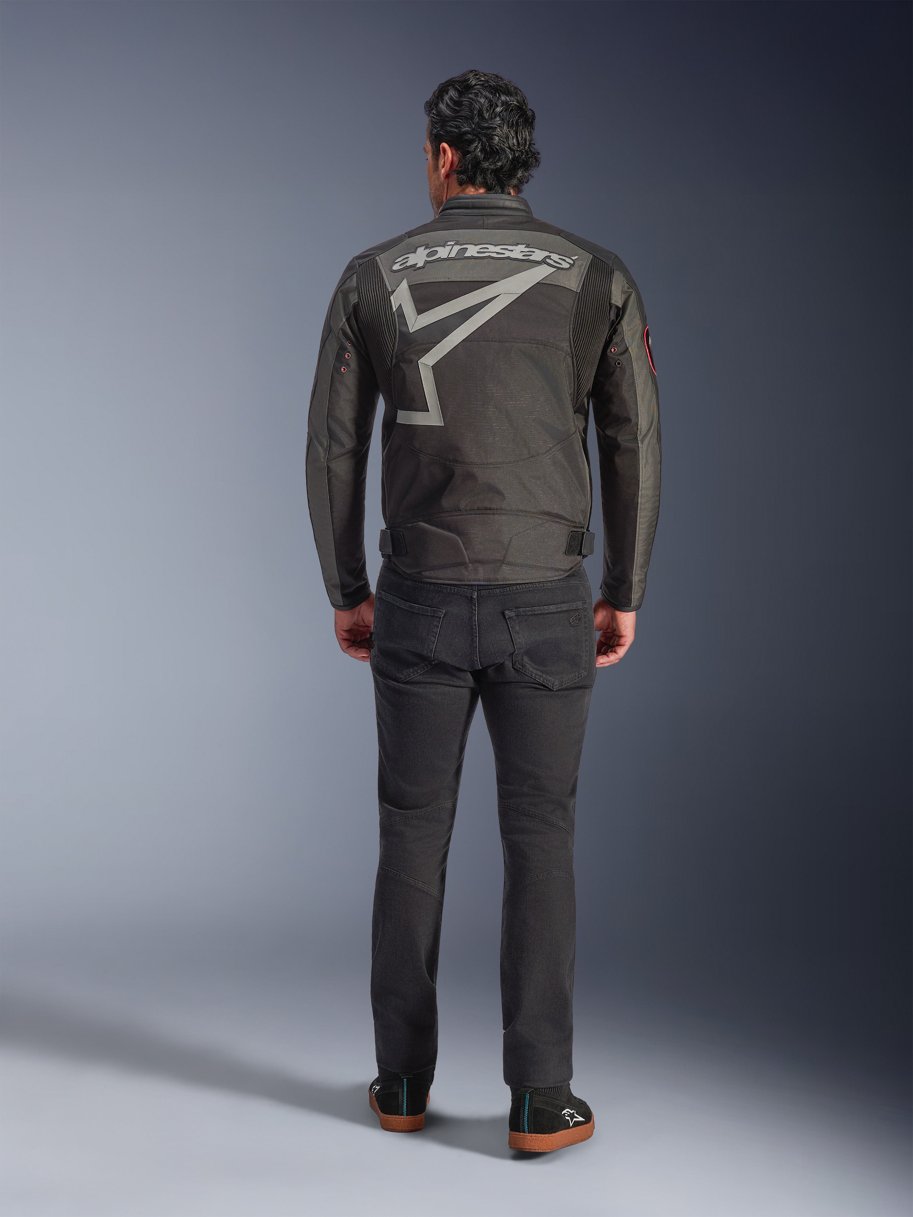 Veste Alpinestars Axton, veste de moto, Noir Anthracite Rouge Tangerine, vue arrière montrant un grand logo Alpinestars gris et un graphisme d'étoile, panneaux accordéon aux épaules, portée avec un pantalon noir et des chaussures noires décontractées.