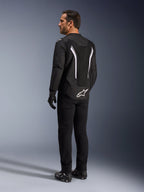 Alpinestars AST V3 Air Jacke, schwarz-weiße Motorradjacke, Rückansicht am Modell, mit Mesheinsätzen für Luftzirkulation und weißem Logo am unteren Rücken, kombiniert mit schwarzer technischer Fahrerausrüstung