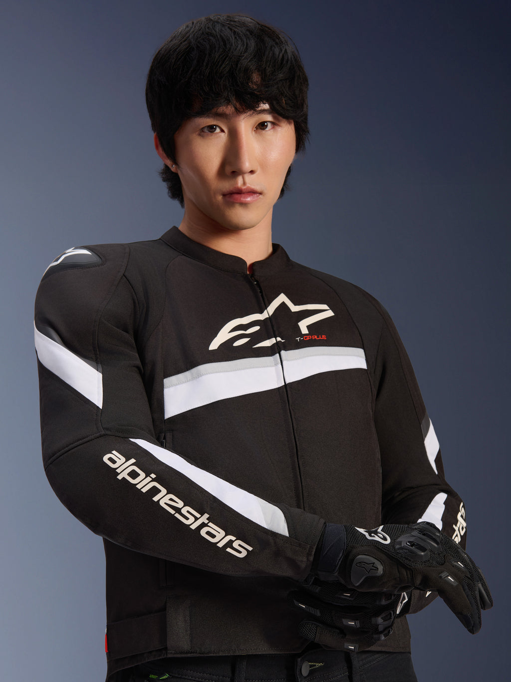 Veste Alpinestars T-GP Plus R V4, noir et blanc, portée par un mannequin homme, veste de moto en textile avec des rayures blanches sur la poitrine et les manches, logo Alpinestars sur la poitrine, associée à des gants de moto noirs