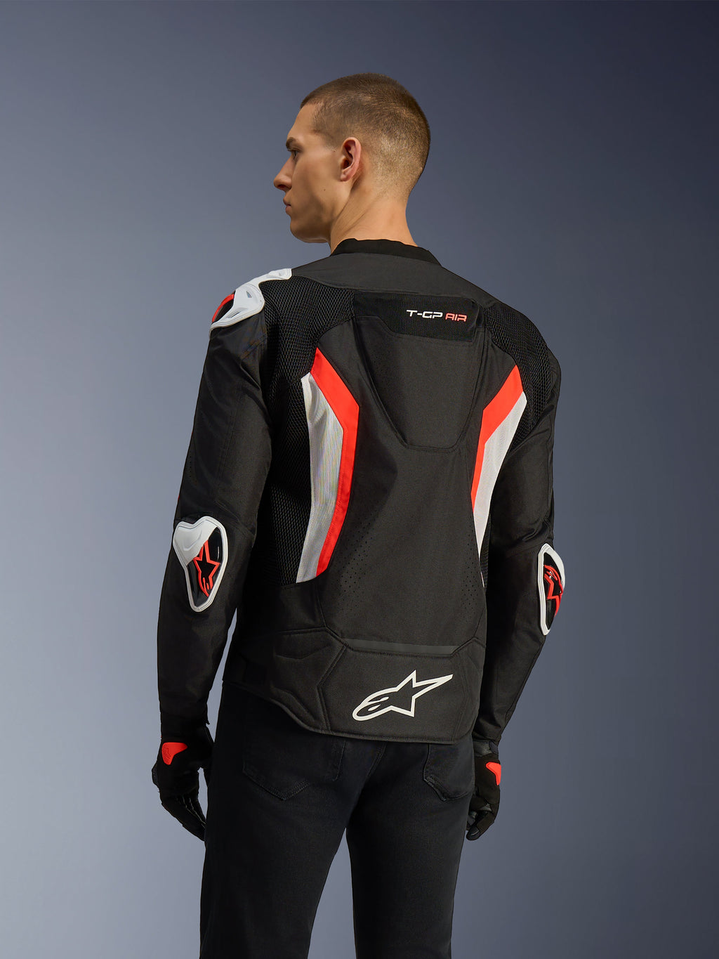 Veste Alpinestars T-GP Air en Noir Rouge Fluo, vue de trois-quarts arrière sur un mannequin, mettant en évidence la bosse aérodynamique, les protections externes Racing GP DFS aux épaules et aux coudes, les panneaux en mesh perforés au laser et les accents blancs et rouge fluo, veste de moto textile à coupe sport.