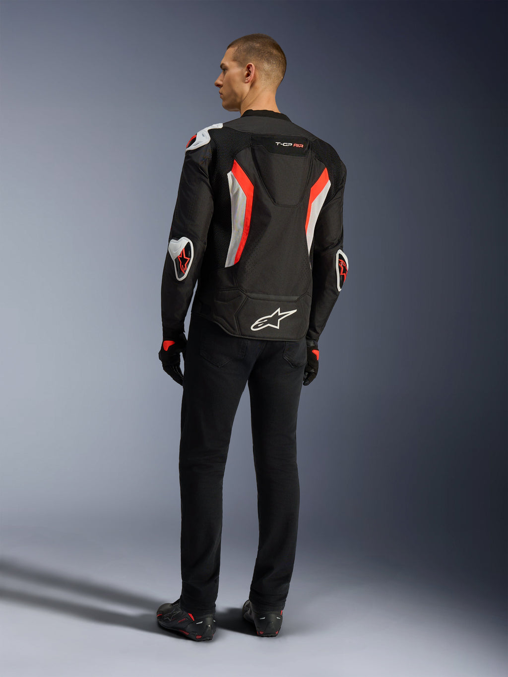Veste Alpinestars T-GP Air, blouson de moto noir rouge fluo, vue arrière sur un modèle, avec panneaux de ventilation en maille, bosse aérodynamique et protections externes blanches aux coudes et aux épaules, porté avec un pantalon noir et des chaussures de moto