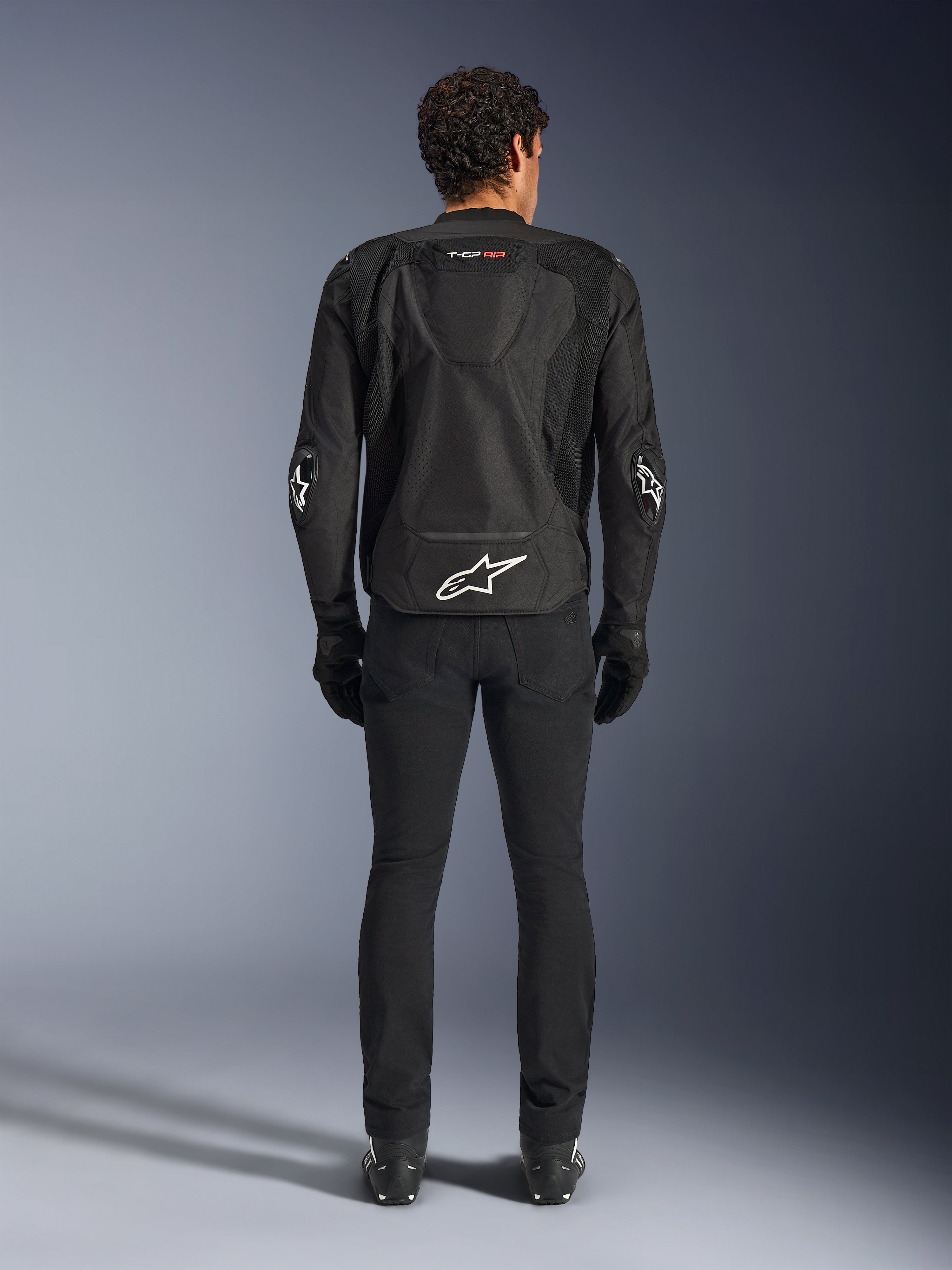 Alpinestars T-GP Air Jacket, noir, vue arrière d'un homme portant la veste de moto textile, présentant des panneaux de ventilation en maille, une bosse aérodynamique et des logos Alpinestars blancs sur le bas du dos et les coudes, portée avec un pantalon et des gants de moto noirs