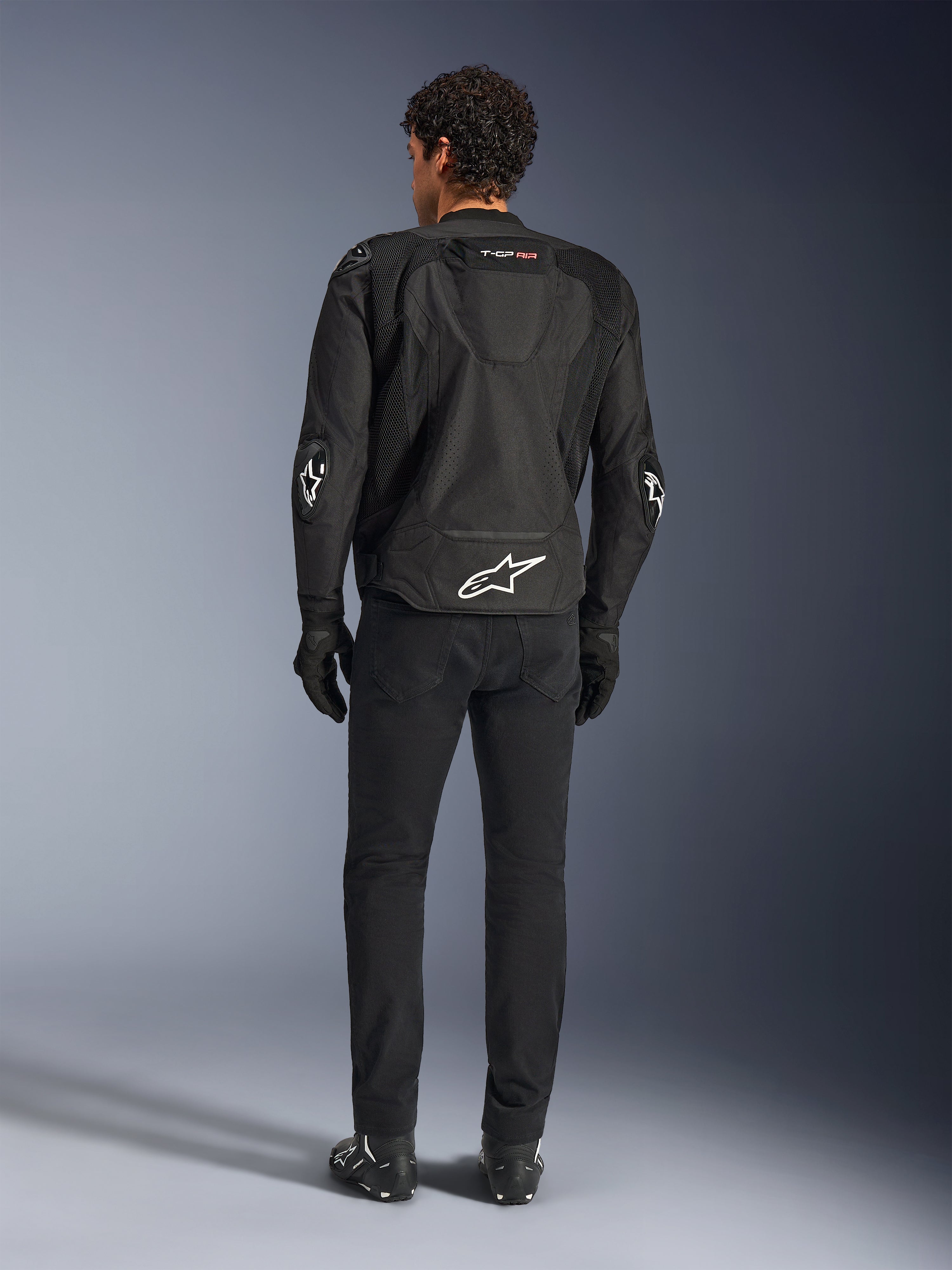 Blouson Alpinestars T-GP Air, noir, vue arrière sur modèle, montrant les panneaux de ventilation en maille 3D et la bosse aérodynamique, avec logo étoile blanc dans le bas du dos et aux coudes, porté avec un pantalon de moto et des gants noirs, équipement de conduite sportive