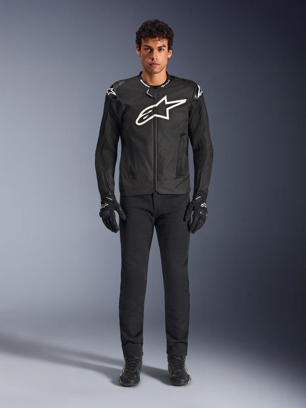 Veste Alpinestars T-GP Air, veste de moto en textile noir avec un grand logo Astars blanc sur la poitrine, portée par un mannequin avec un pantalon et des gants de moto noirs, dotée de panneaux perforés et d'une ventilation en mesh pour la conduite sportive