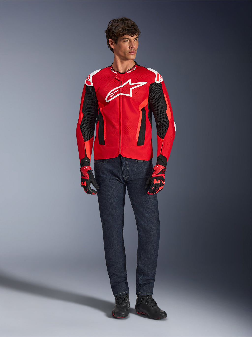 Alpinestars T-GP Air Jacke, Hellrot Fluo-Rot Schwarz, männliches Model in kompletter Motorradausrüstung mit dunkelblauer Jeans und passenden Handschuhen, Sport-Textiljacke mit perforierten Mesh-Einsätzen und weißem Astars-Logo, Studioaufnahme.