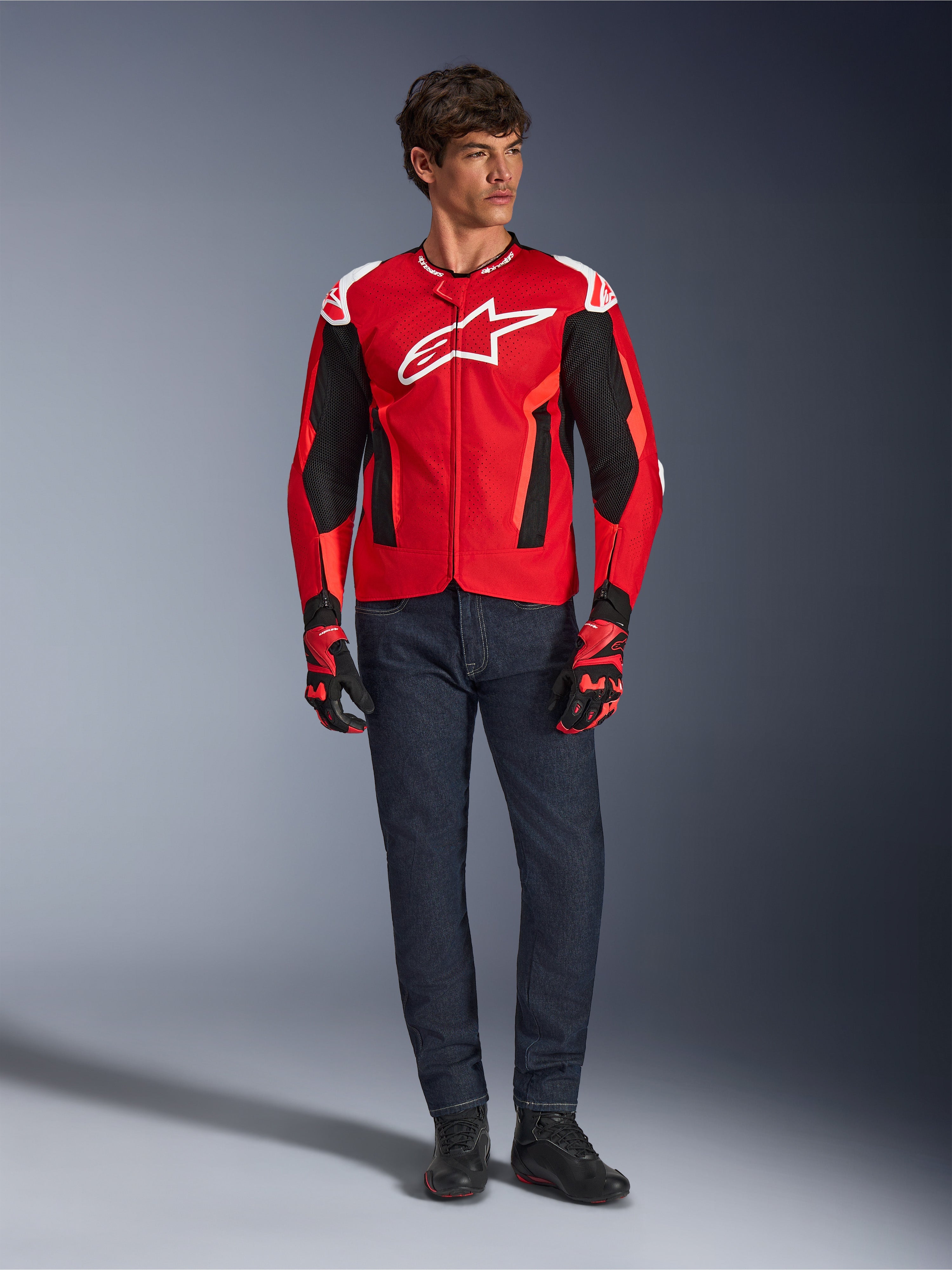 Alpinestars T-GP Air Jacke, Hellrot Fluo-Rot Schwarz, männliches Model in kompletter Motorradausrüstung mit dunkelblauer Jeans und passenden Handschuhen, Sport-Textiljacke mit perforierten Mesh-Einsätzen und weißem Astars-Logo, Studioaufnahme.