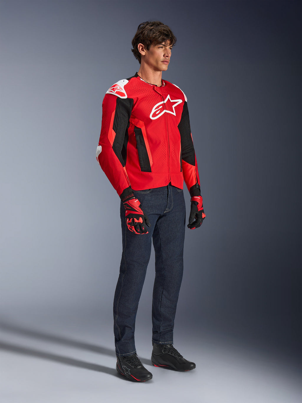 Alpinestars T-GP Air Jacke, sportliche Motorradjacke, leuchtendes Rot und Schwarz mit weißen Akzenten, getragen von einem Model mit dunkelblauen Jeans und roten Rennhandschuhen, mit High-Flow-Mesh-Einsätzen und weißen Schulterschleifern, aerodynamische Sport-Passform für Fahrten bei warmem Wetter.