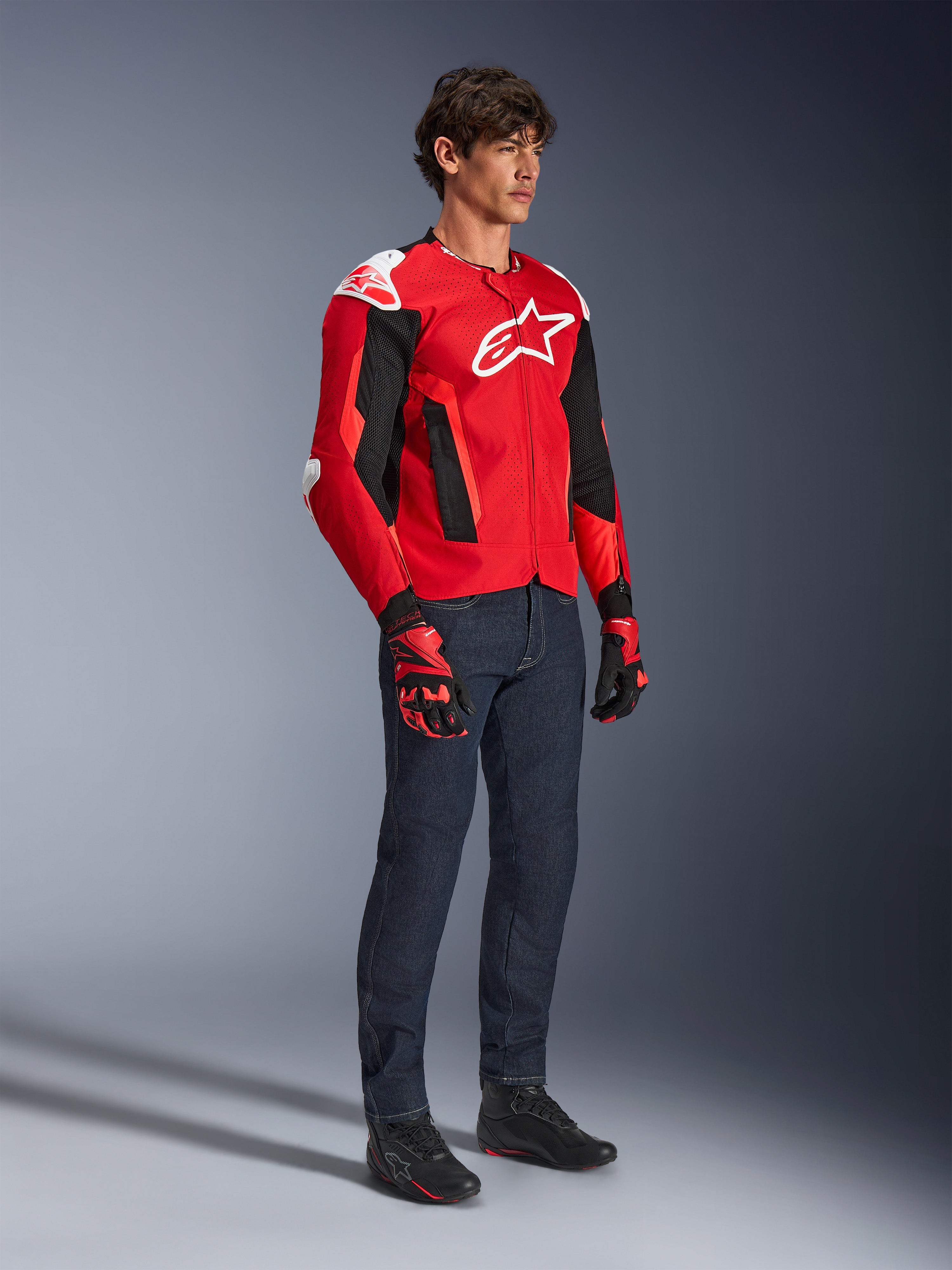 Alpinestars T-GP Air Jacke, sportliche Motorradjacke, leuchtendes Rot und Schwarz mit weißen Akzenten, getragen von einem Model mit dunkelblauen Jeans und roten Rennhandschuhen, mit High-Flow-Mesh-Einsätzen und weißen Schulterschleifern, aerodynamische Sport-Passform für Fahrten bei warmem Wetter.