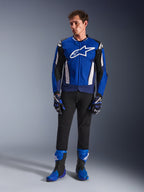 Modèle masculin portant la veste Alpinestars T-GP Air en bleu vif, bleu marine et blanc, associée à un pantalon noir, des gants de course assortis et des bottes de moto bleues et noires, photo de studio présentant une tenue de route sportive complète.