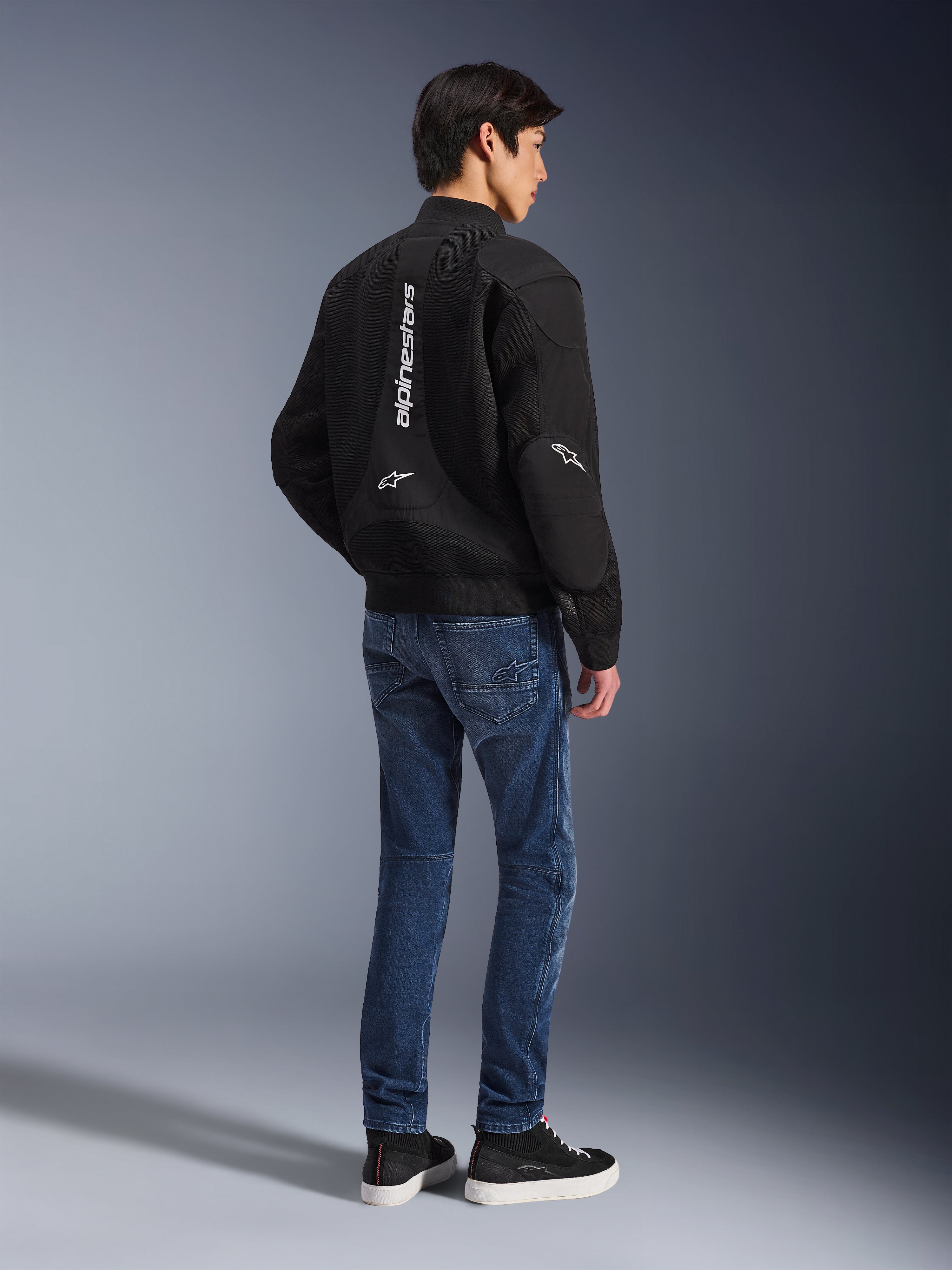 Model in der Alpinestars Flight Air Jacke in Schwarz, Rückansicht, kombiniert mit blauen Motorradjeans und schwarzen Sneakern, mit vertikalem weißem Alpinestars-Schriftzug und 3D-Mesh-Belüftungseinsätzen.