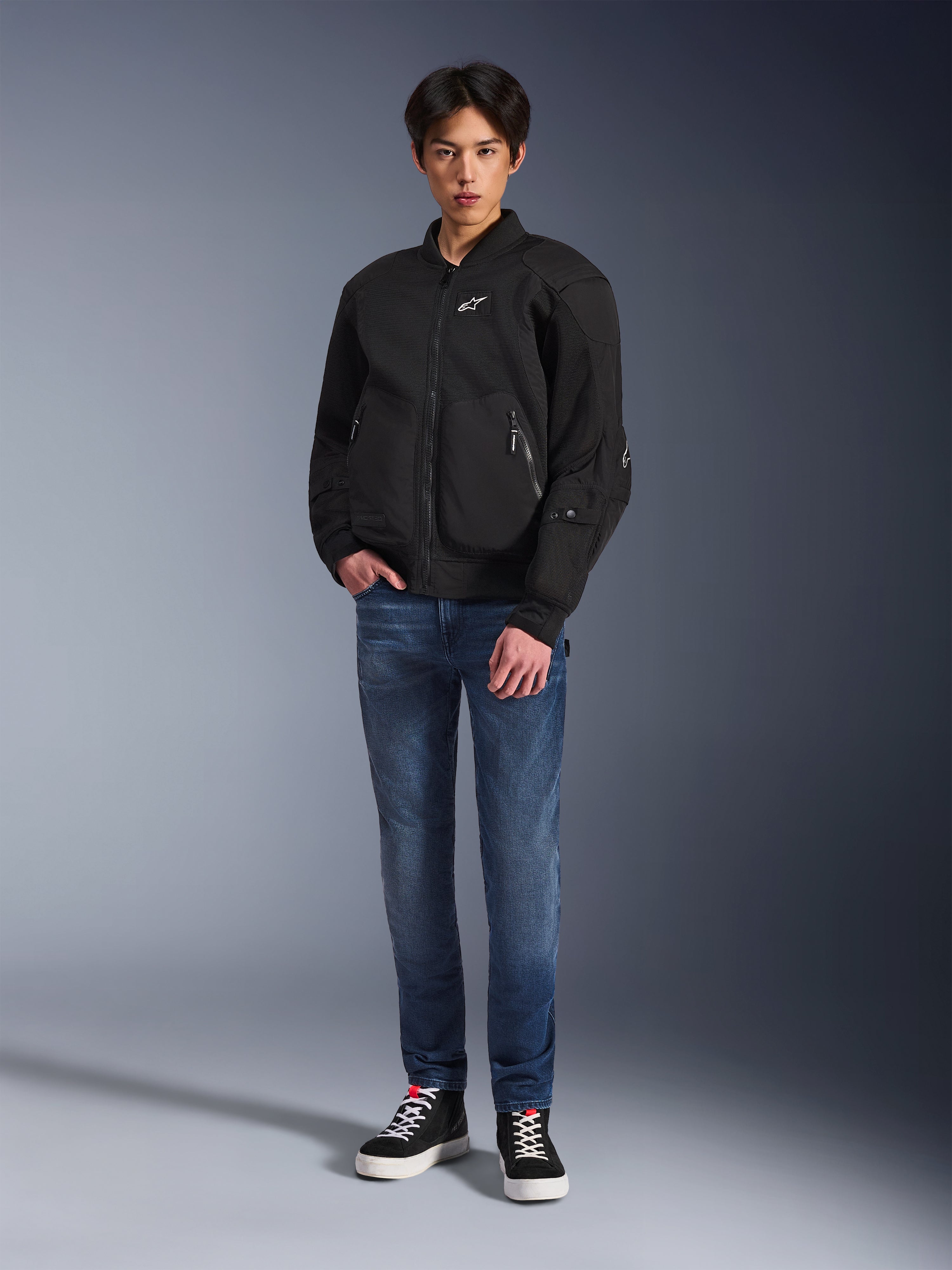 Model in der Alpinestars Flight Air Jacke in Schwarz, eine urbane Motorradjacke im Bomber-Stil mit 3D-Mesh-Einsätzen und Frontreißverschluss, kombiniert mit blauen Jeans und schwarzen Sneakern, moderne Streetwear-inspirierte Motorradbekleidung.