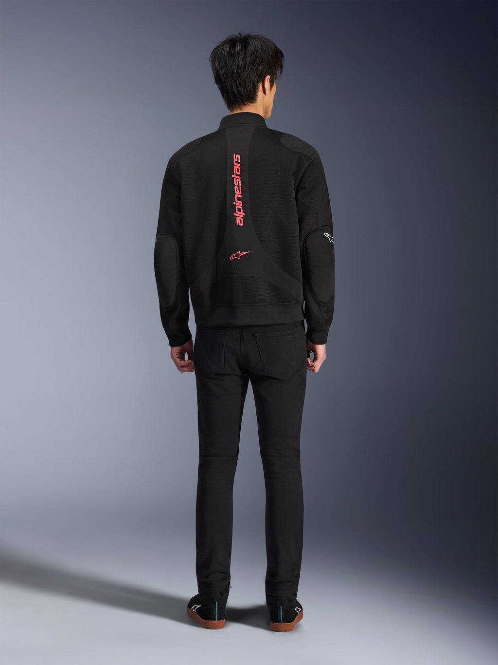 Veste Alpinestars Flight Air en Noir et Rouge Vif, vue arrière d'une veste de moto de style bomber urbain, arborant le lettrage et le logo Alpinestars verticaux rouges sur le dos, conception légère en maille 3D, portée avec un pantalon noir pour un look contemporain.