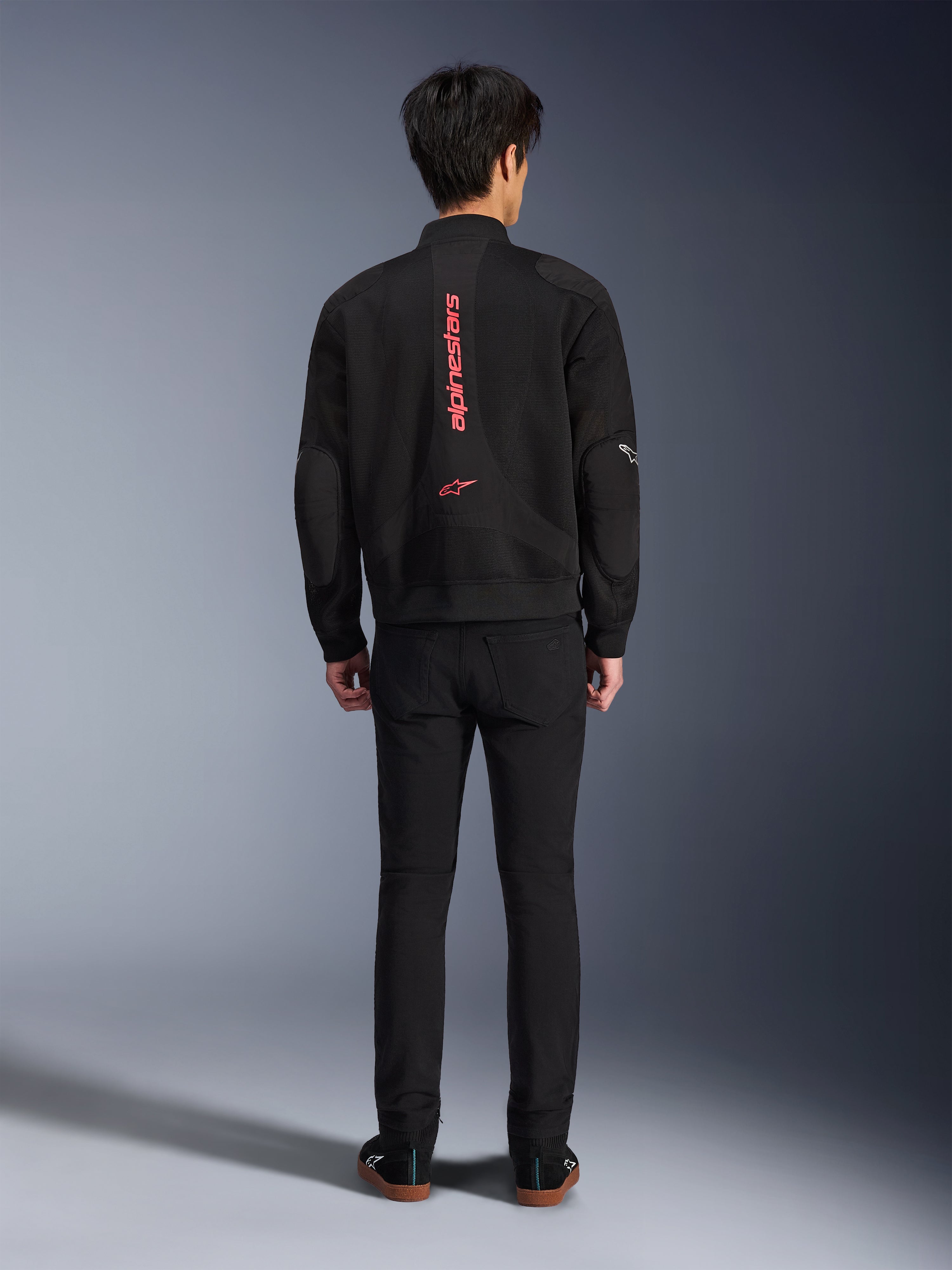 Veste Alpinestars Flight Air en Noir et Rouge Vif, vue arrière d'une veste de moto de style bomber urbain, arborant le lettrage et le logo Alpinestars verticaux rouges sur le dos, conception légère en maille 3D, portée avec un pantalon noir pour un look contemporain.