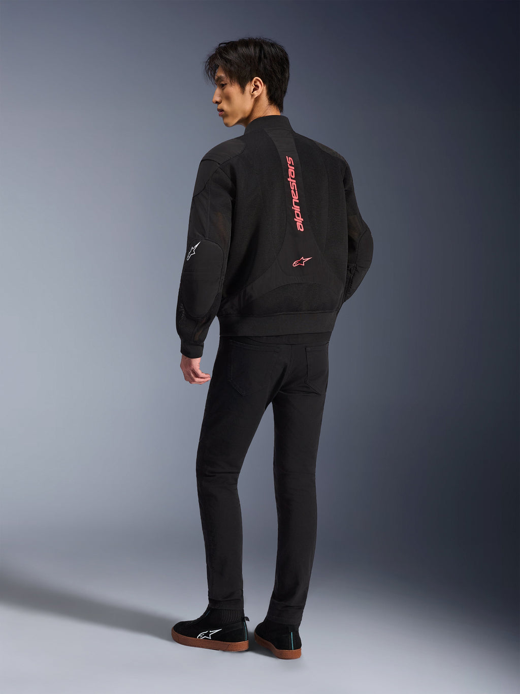 Modèle masculin portant la veste Alpinestars Flight Air en noir et rouge vif, vue arrière montrant le marquage vertical rouge au dos et la construction en maille 3D, associée à un pantalon noir et des baskets, style moto urbain