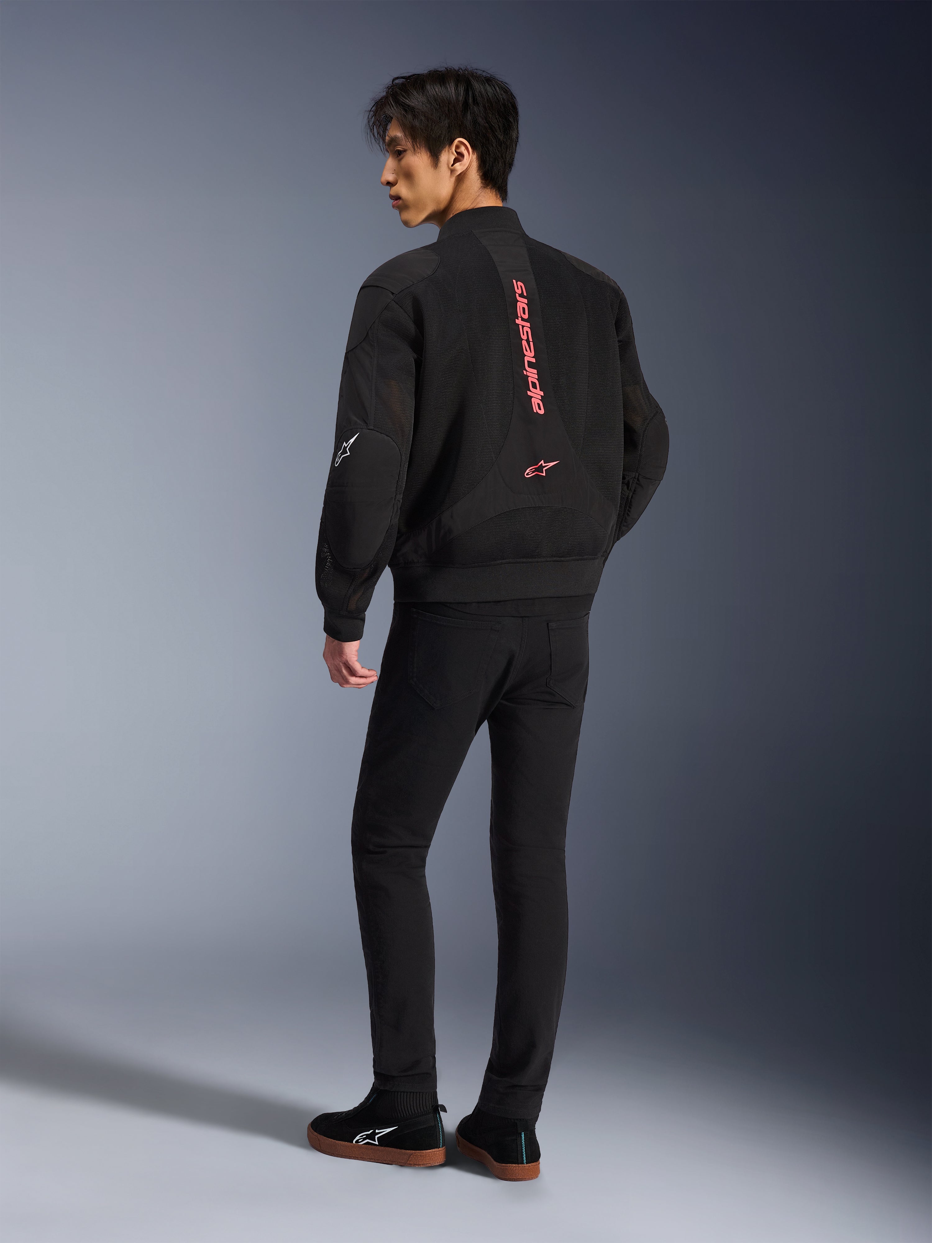 Modèle masculin portant la veste Alpinestars Flight Air en noir et rouge vif, vue arrière montrant le marquage vertical rouge au dos et la construction en maille 3D, associée à un pantalon noir et des baskets, style moto urbain