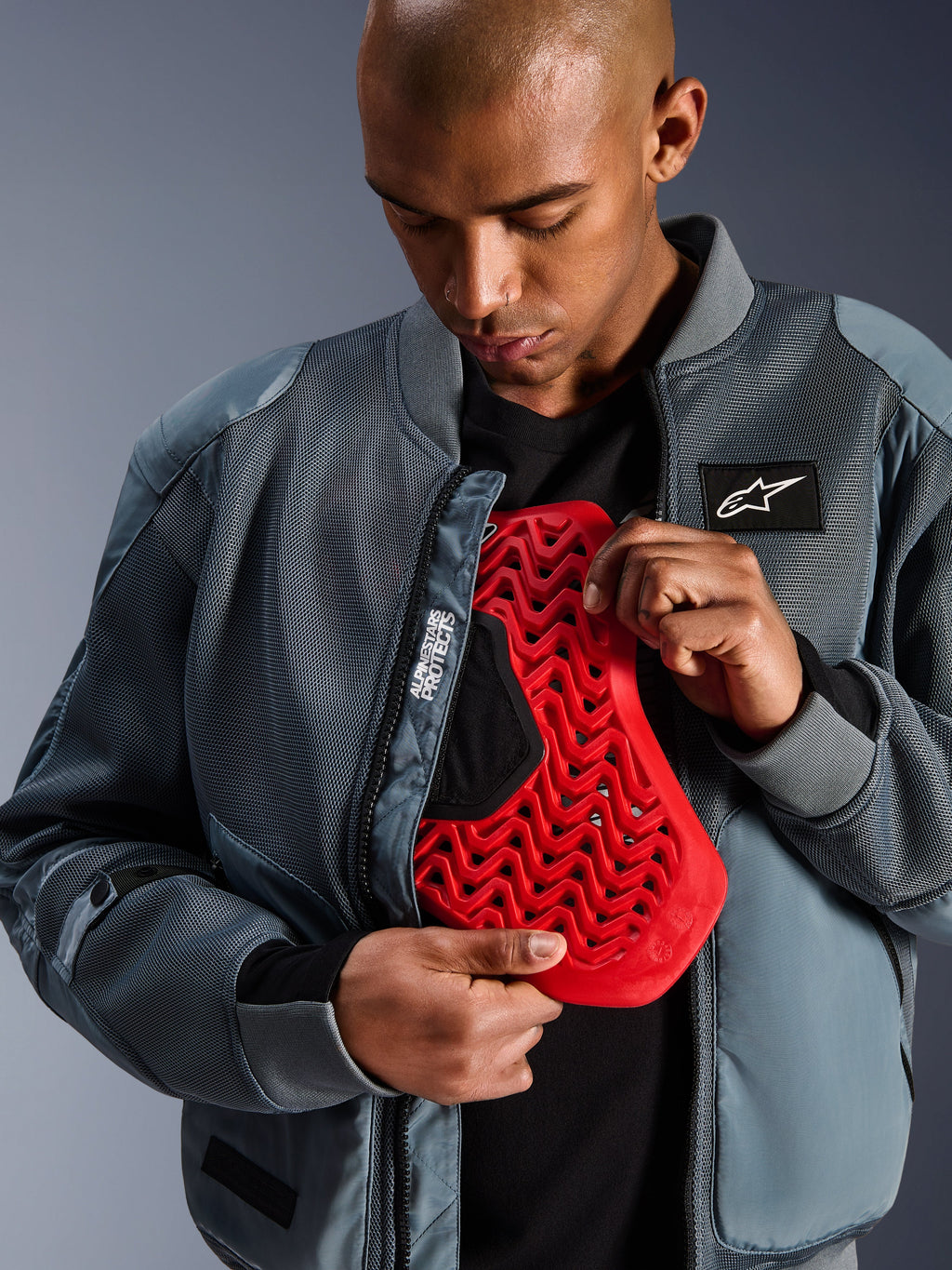 Alpinestars Flight Air Jacke in Storm Gray, urbane Motorrad-Bomberjacke mit 3D-Mesh-Paneelen für Belüftung, geöffnet mit sichtbarem rotem Nucleon Rückenprotektor-Einsatz, Alpinestars Protects Branding und moderne Streetwear-Ästhetik