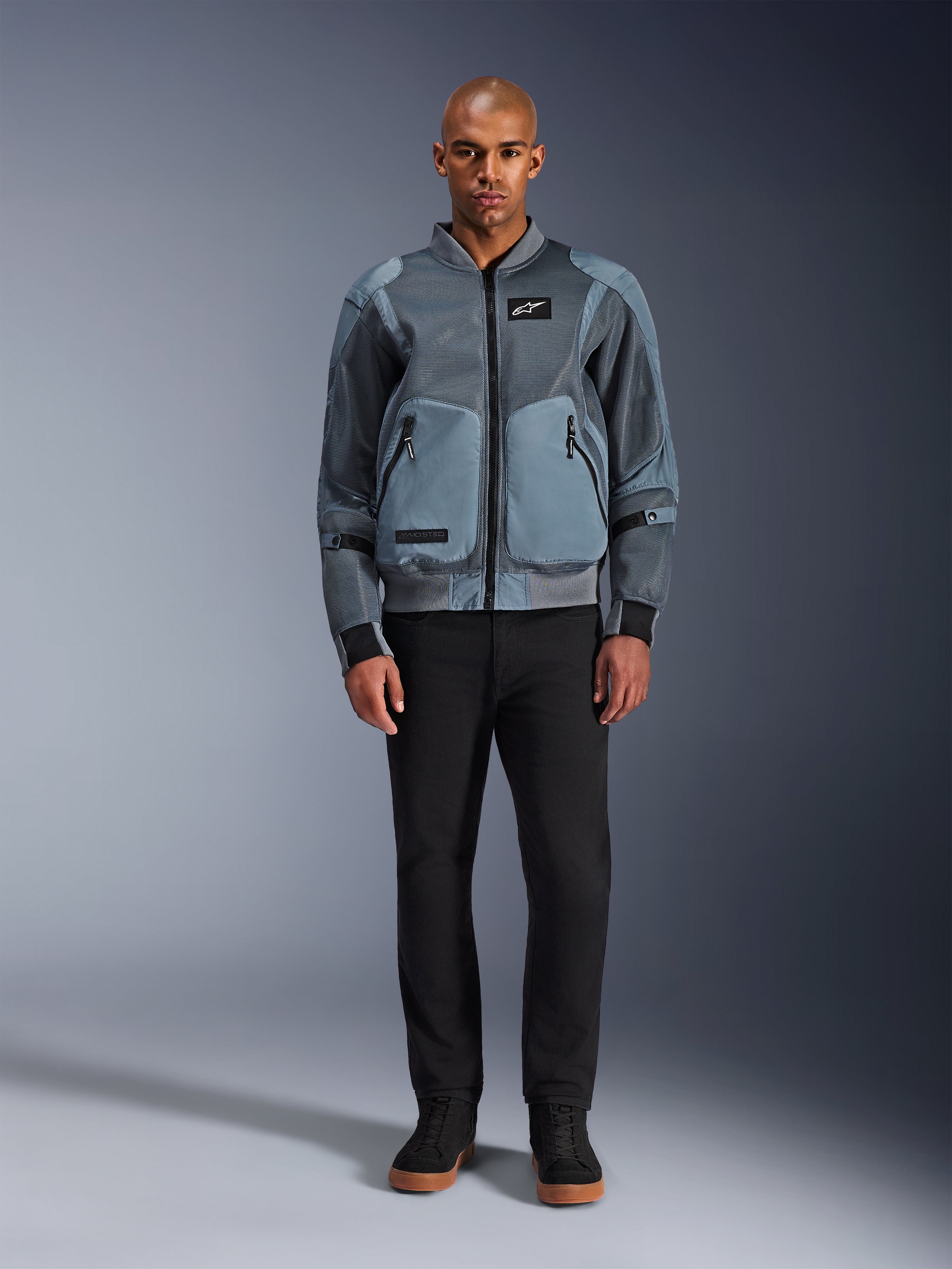 Alpinestars Flight Air Jacke in Storm Gray, urban inspirierte Bomber-Motorradjacke mit 3D-Mesh-Einsätzen und robusten Fronttaschen, getragen von einem Mann mit schwarzer Hose und schwarzen High-Top-Sneakern, moderne Motorradbekleidung im Streetstyle