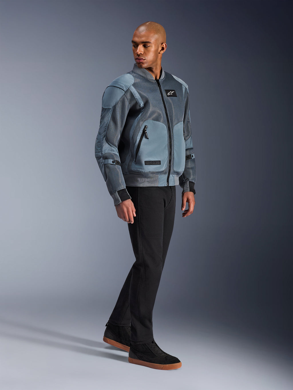 Alpinestars Flight Air Jacke, Storm Gray, urbane Motorradjacke im Bomber-Stil, getragen von einem männlichen Model mit schwarzer Hose und Sneakern, mit großflächigen 3D-Mesh-Belüftungseinsätzen und externen Protektorentaschen, moderne Streetwear-Schutzkleidung für Fahrten bei heißem Wetter