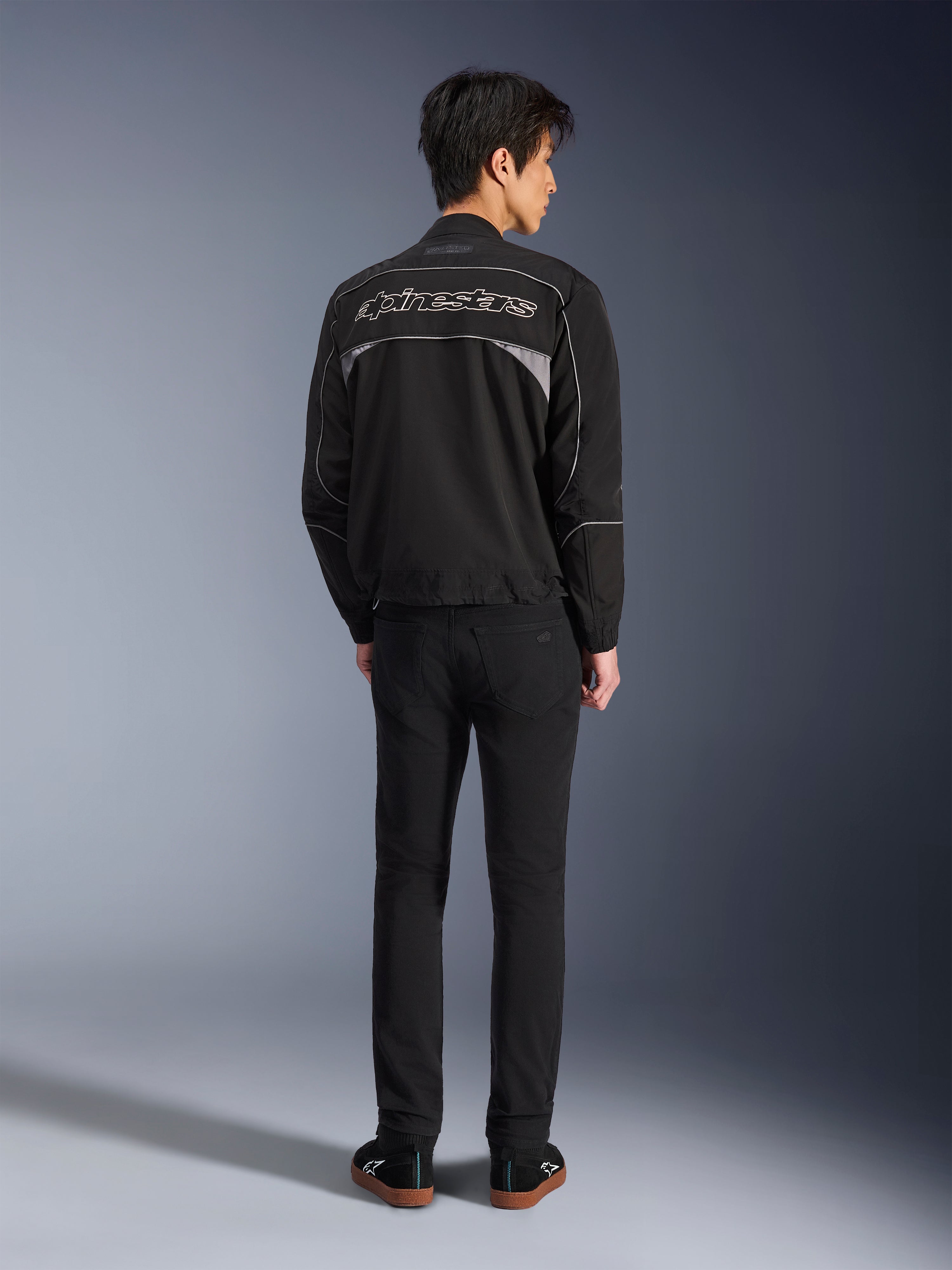 Alpinestars Unite Jacke, Motorradjacke in Schwarz und Dunkelgrau, Rückansicht mit Alpinestars-Umrisslogo auf dem Rücken, graue reflektierende Einsätze, minimalistisches urbanes Design, getragen mit schwarzer Hose und schwarzen Schuhen.
