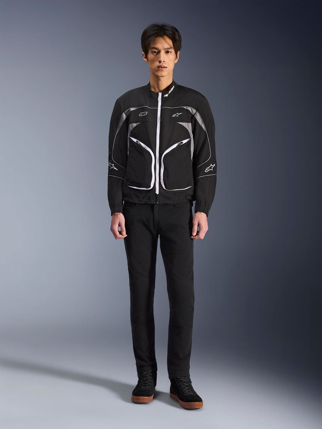 Alpinestars Unite Jacke, Motorradjacke, Schwarz und Dunkelgrau, getragen von einem Model mit schwarzer Hose und schwarzen Stiefeln, weiße Kontrast-Reißverschlüsse, reflektierende Paspeln, minimalistisches urbanes Design