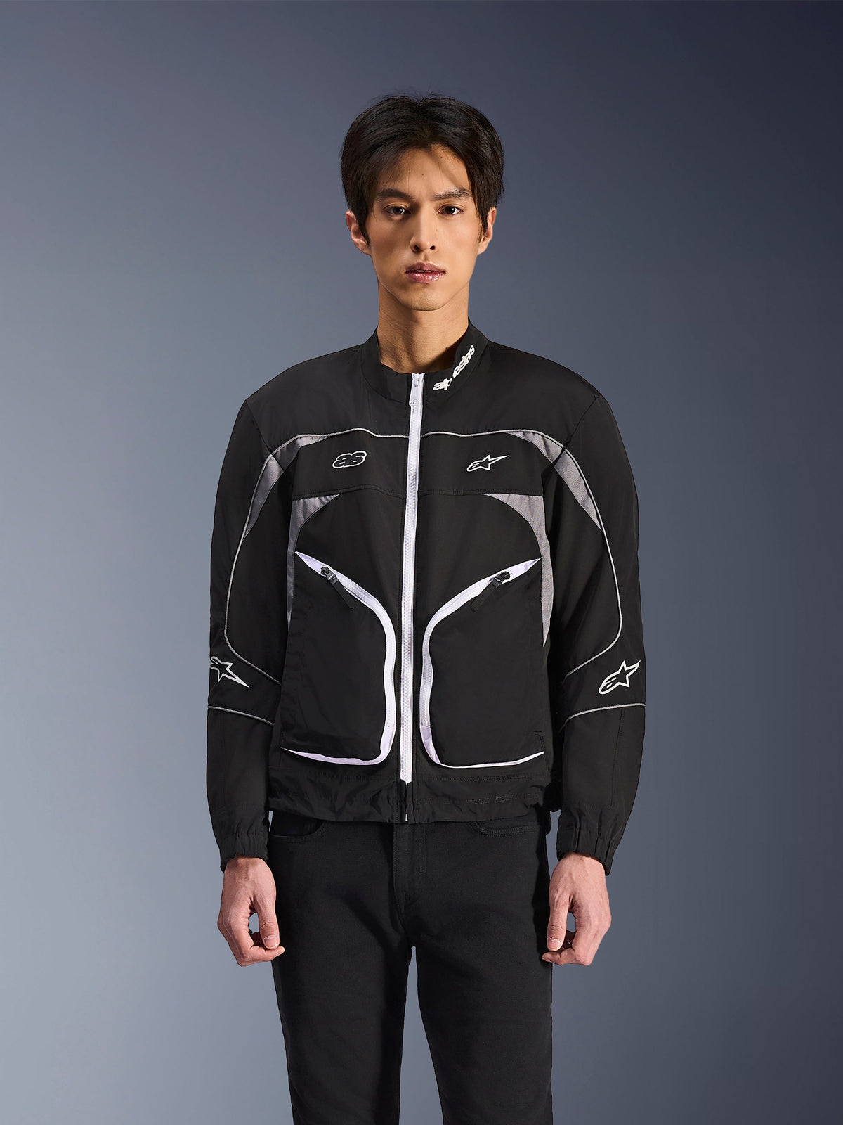 Veste Alpinestars Unite, veste de moto urbaine minimaliste, noir et gris foncé avec des accents blancs, dotée d'une fermeture éclair frontale blanche, de liserés réfléchissants et du logo Alpinestars sur la poitrine et les manches, style streetwear contemporain pour les motards.
