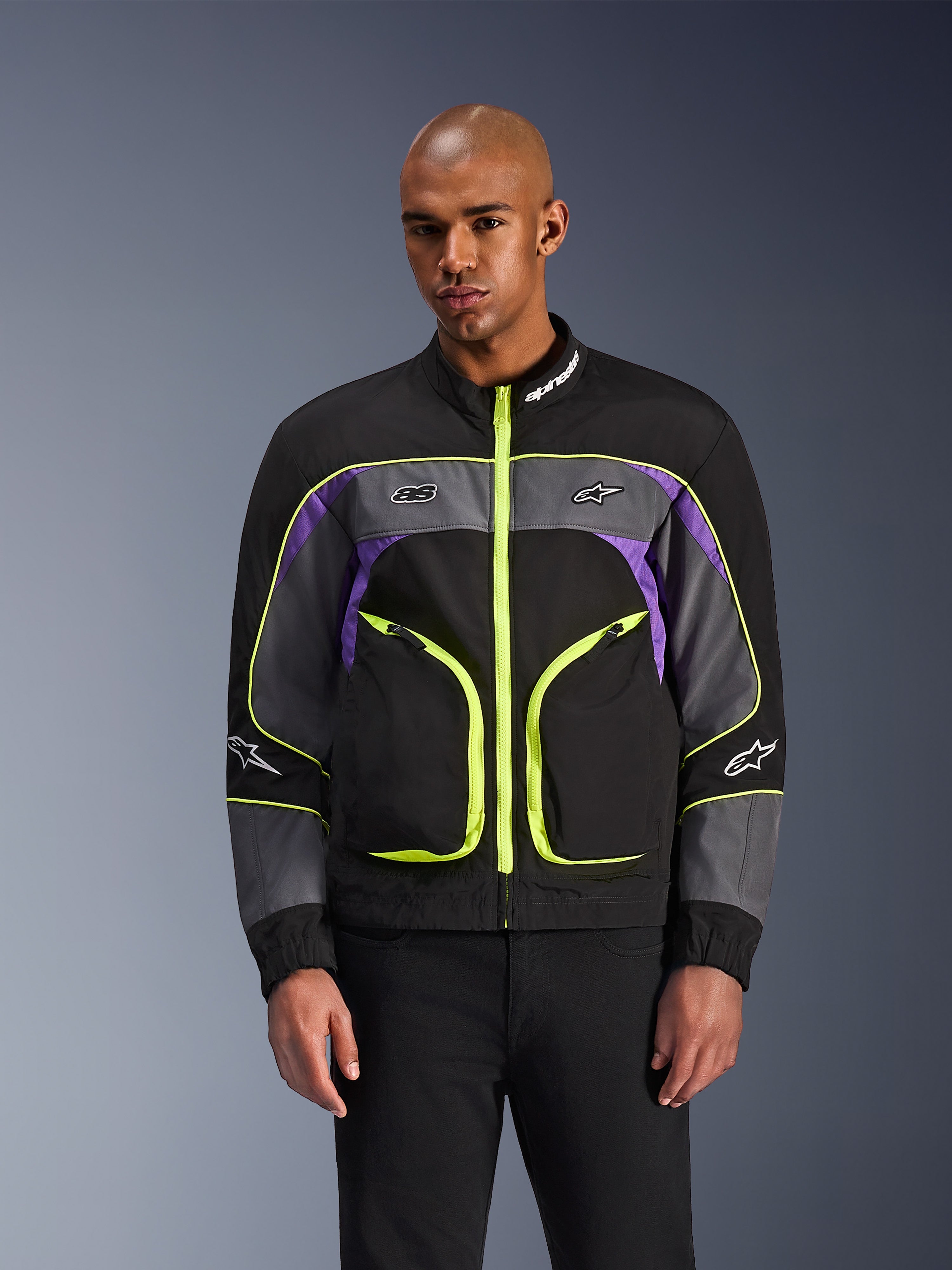 Alpinestars Unite Jacket, veste de moto urbaine, noir asphalte et violet avec accents jaune fluo, dotée de grandes poches avant avec fermetures éclair fluo, col haut avec logo blanc, portée par un mannequin, équipement de protection contemporain inspiré du streetwear