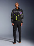 Männliches Model trägt die Alpinestars Unite Jacke in Black Asphalt Violet, mit schwarzer Basis, grauen und lila Einsätzen sowie leuchtend neongelben Reißverschlüssen und Details, kombiniert mit schwarzer Hose und Schuhen, minimalistische urbane Motorradjacke.